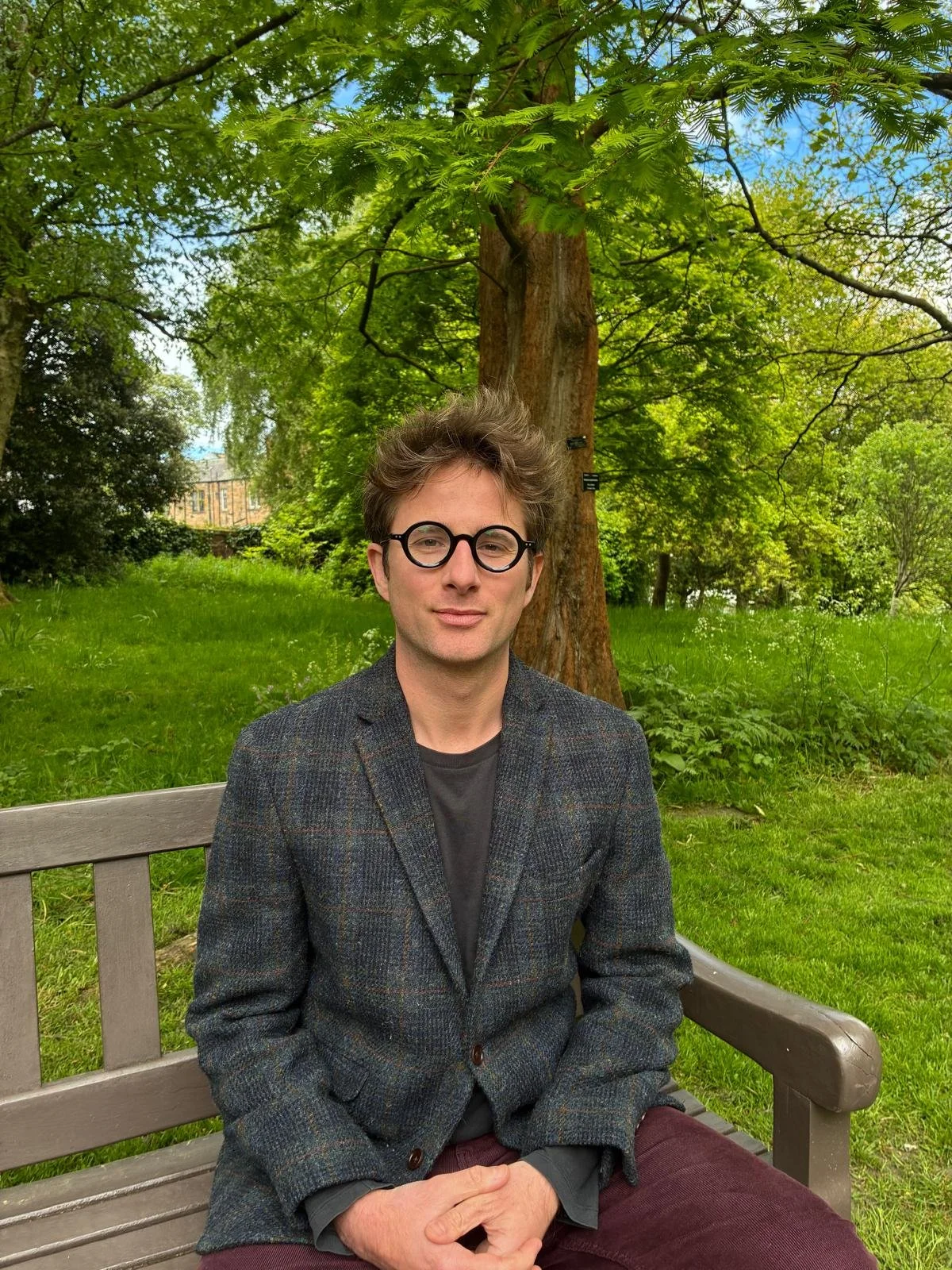 A man with glasses and messy hair, wearing a gray blazer and dark shirt, sitting on a park bench with greenery and trees in the background.