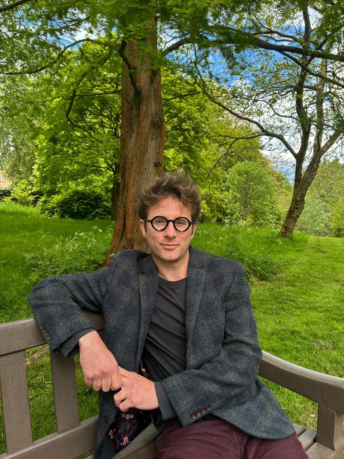 A man with glasses and brown hair sitting on a park bench surrounded by trees and green grass.