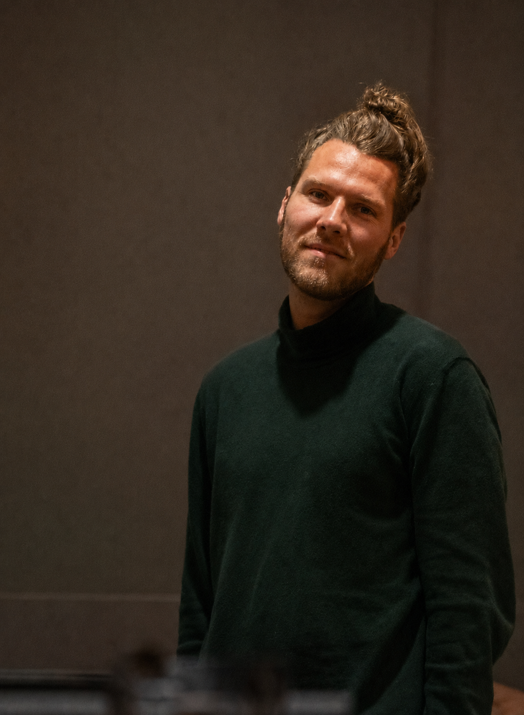 Ein junger Mann mit dunkelblondem hochgestecktem Haar und Bart, er trägt einen schwarzen Pullover, steht vor einem dunklen Hintergrund und lächelt leicht.