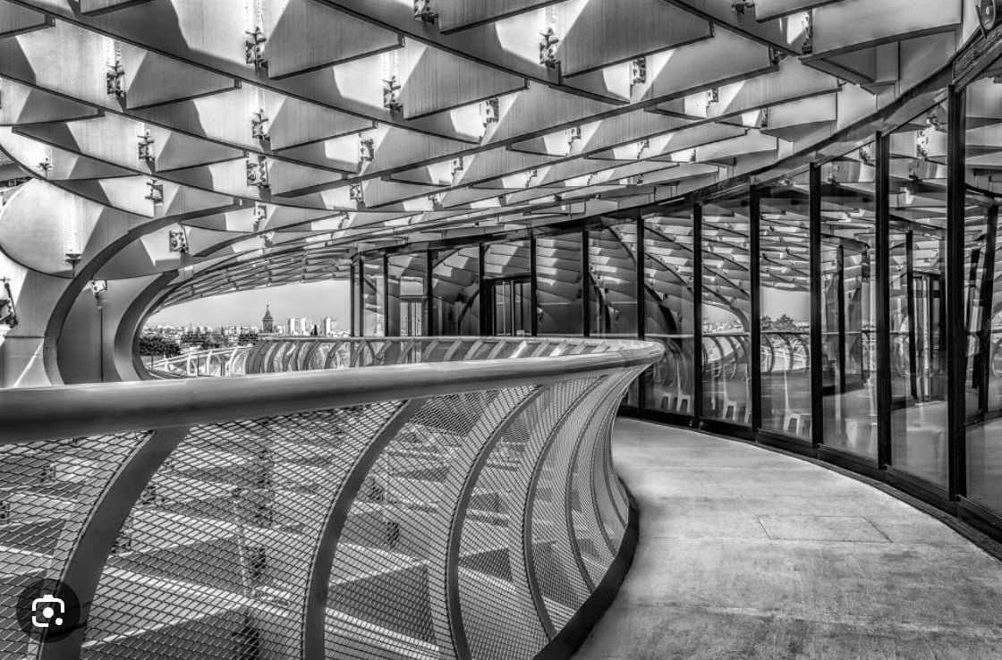 A black and white photo of a curved walkway with a metal railing, under a wooden, geometric ceiling.