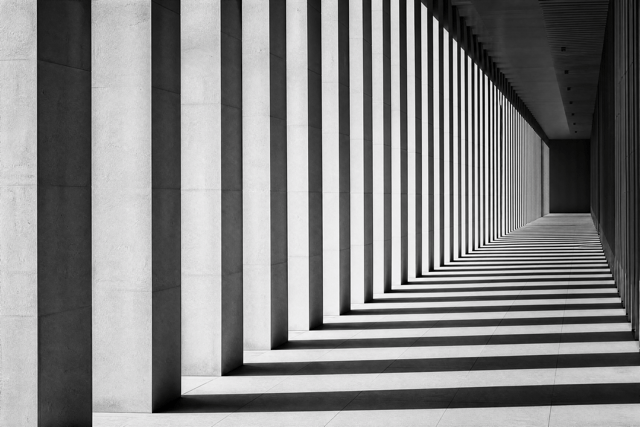 Black and white image of a hallway with strong geometric patterns created by light and shadow from vertical pillars.