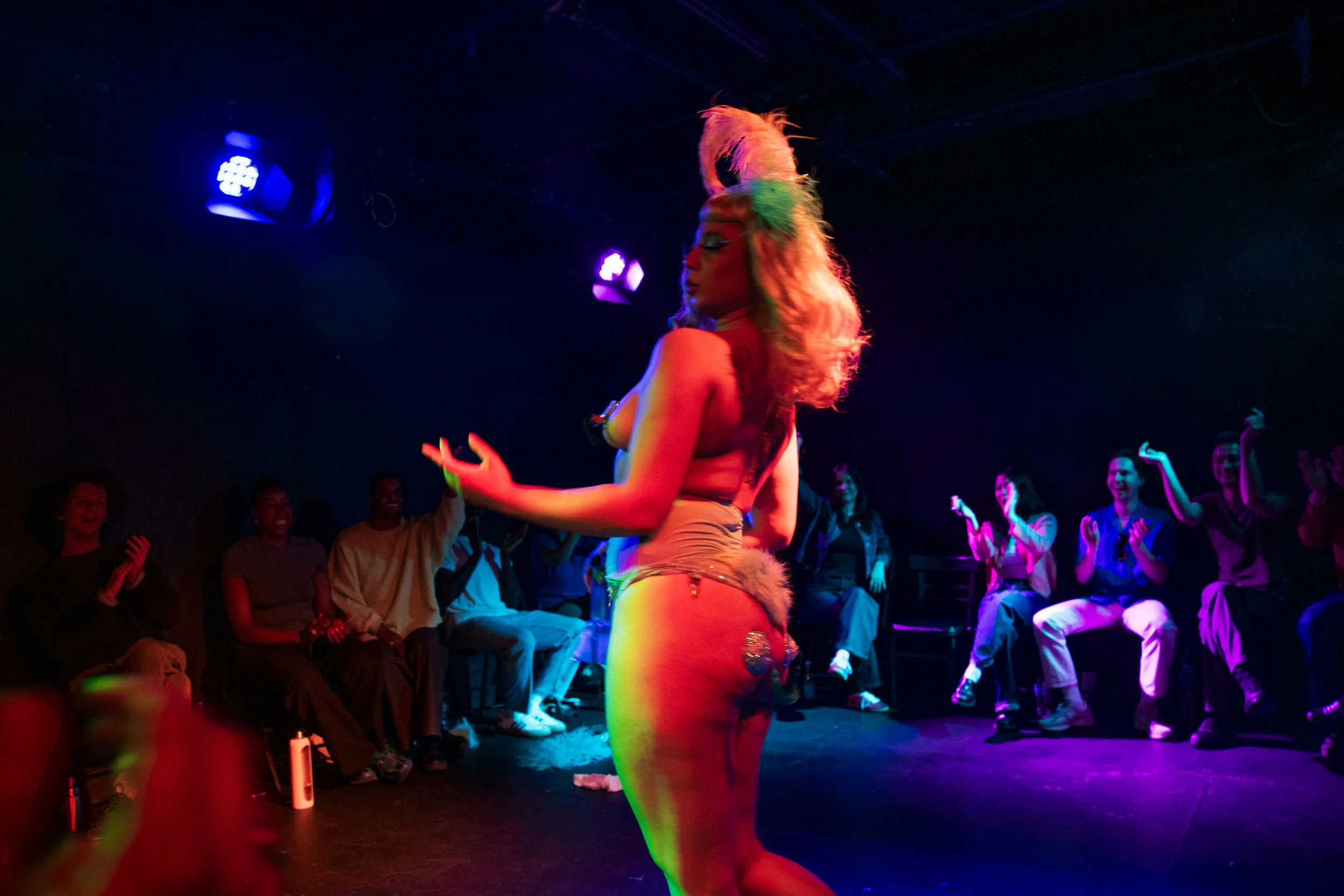 A performer in colorful stage costume, with vibrant hair and makeup, dancing in front of an audience in a darkened theater with blue and purple stage lighting.