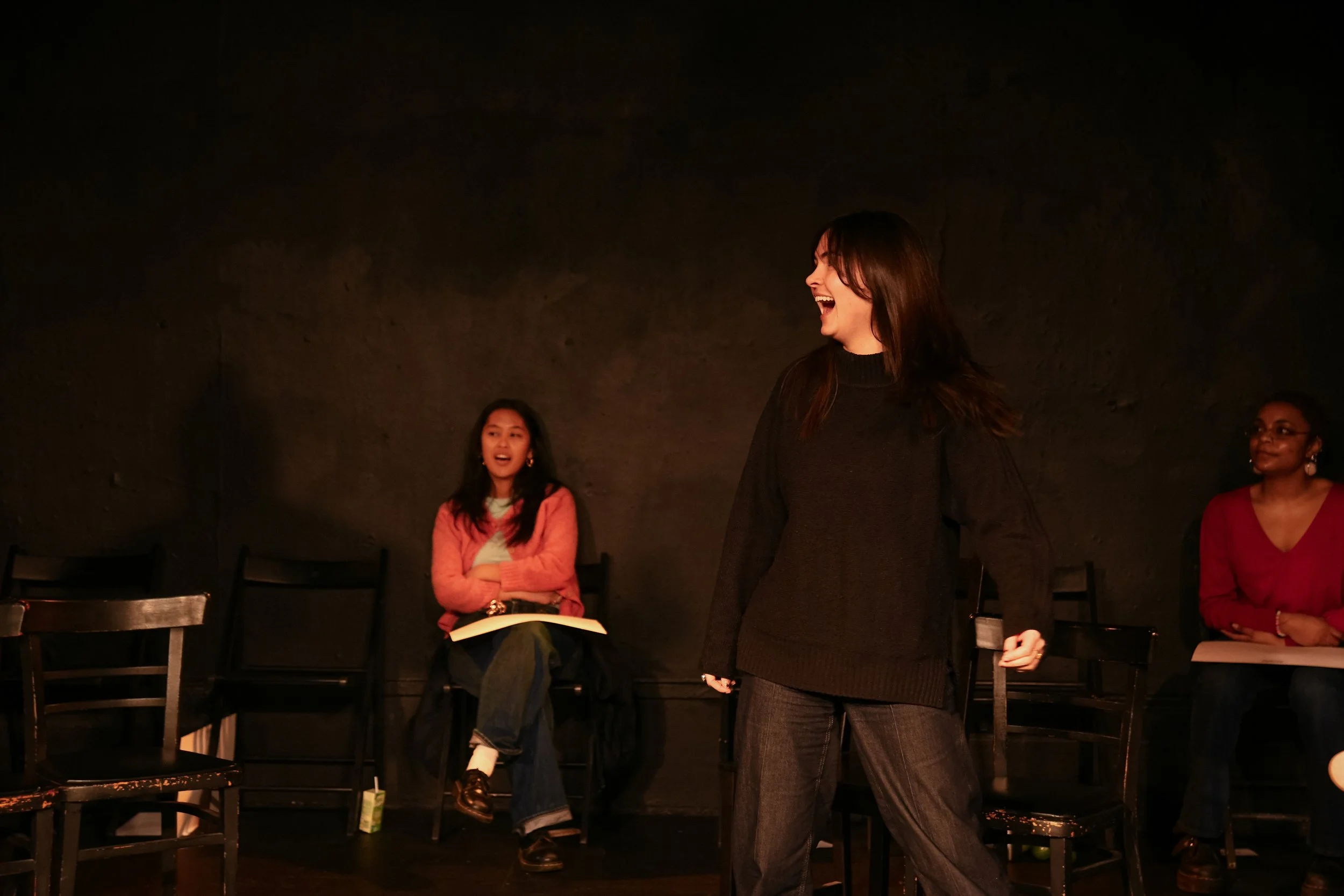 A woman is standing and laughing in front of three seated women in a dark room, possibly during a performance or a conversation, with chairs lined up against a dark wall.