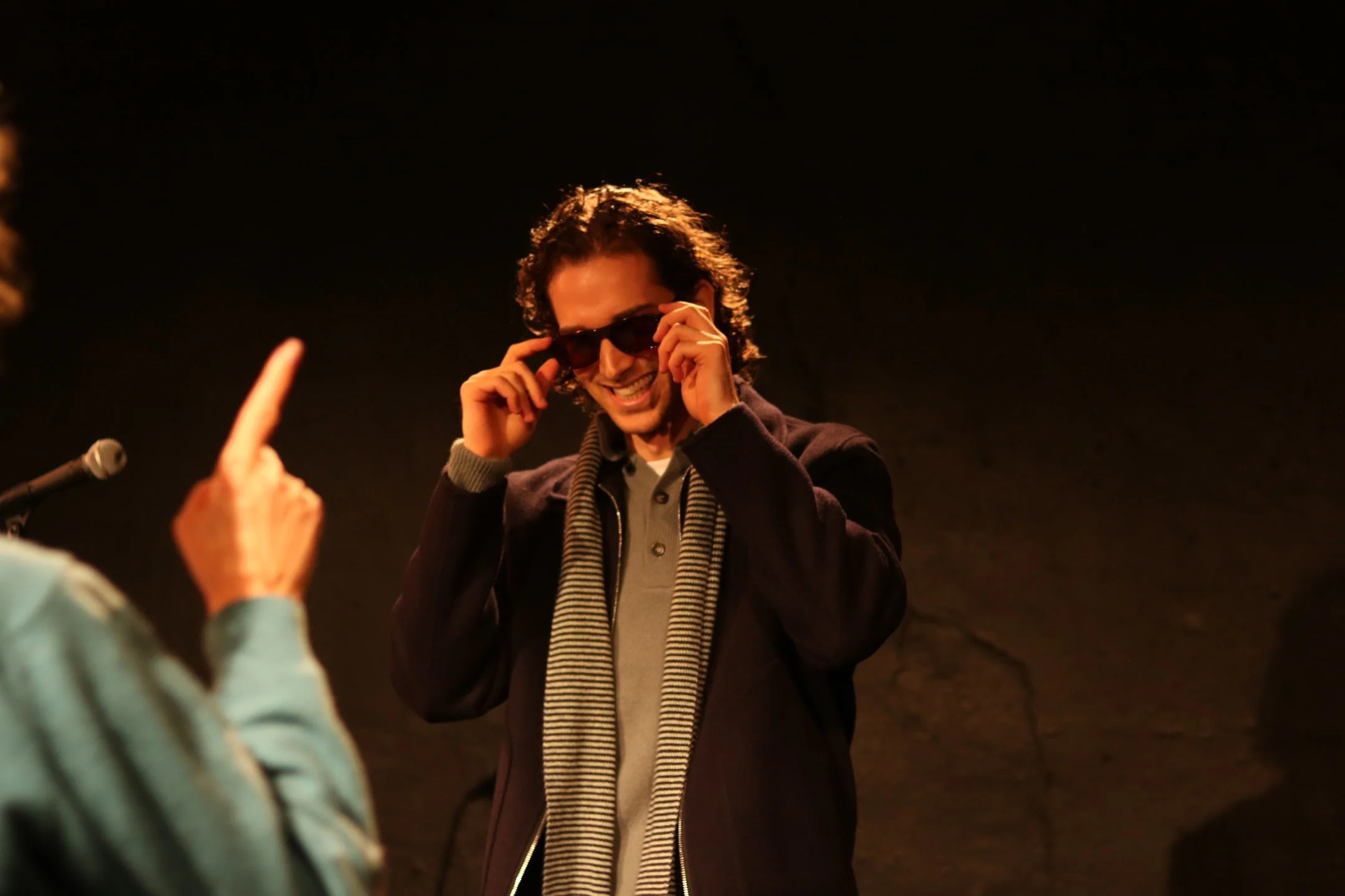 A man with curly hair and glasses smiling while adjusting his sunglasses, wearing a striped scarf and jacket, interacting with someone in the foreground. The background is dark.