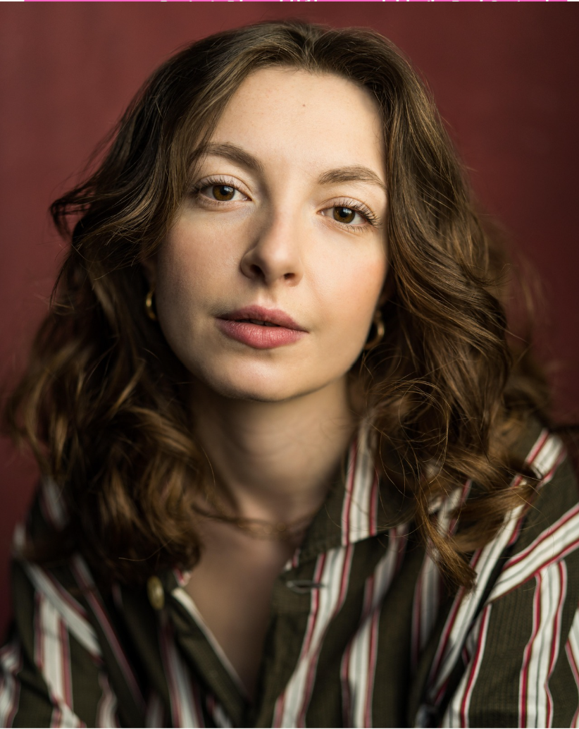 A young woman with wavy brown hair and light skin, wearing gold hoop earrings and a striped button-up shirt, looking directly at the camera with a neutral expression, against a reddish-burgundy background.