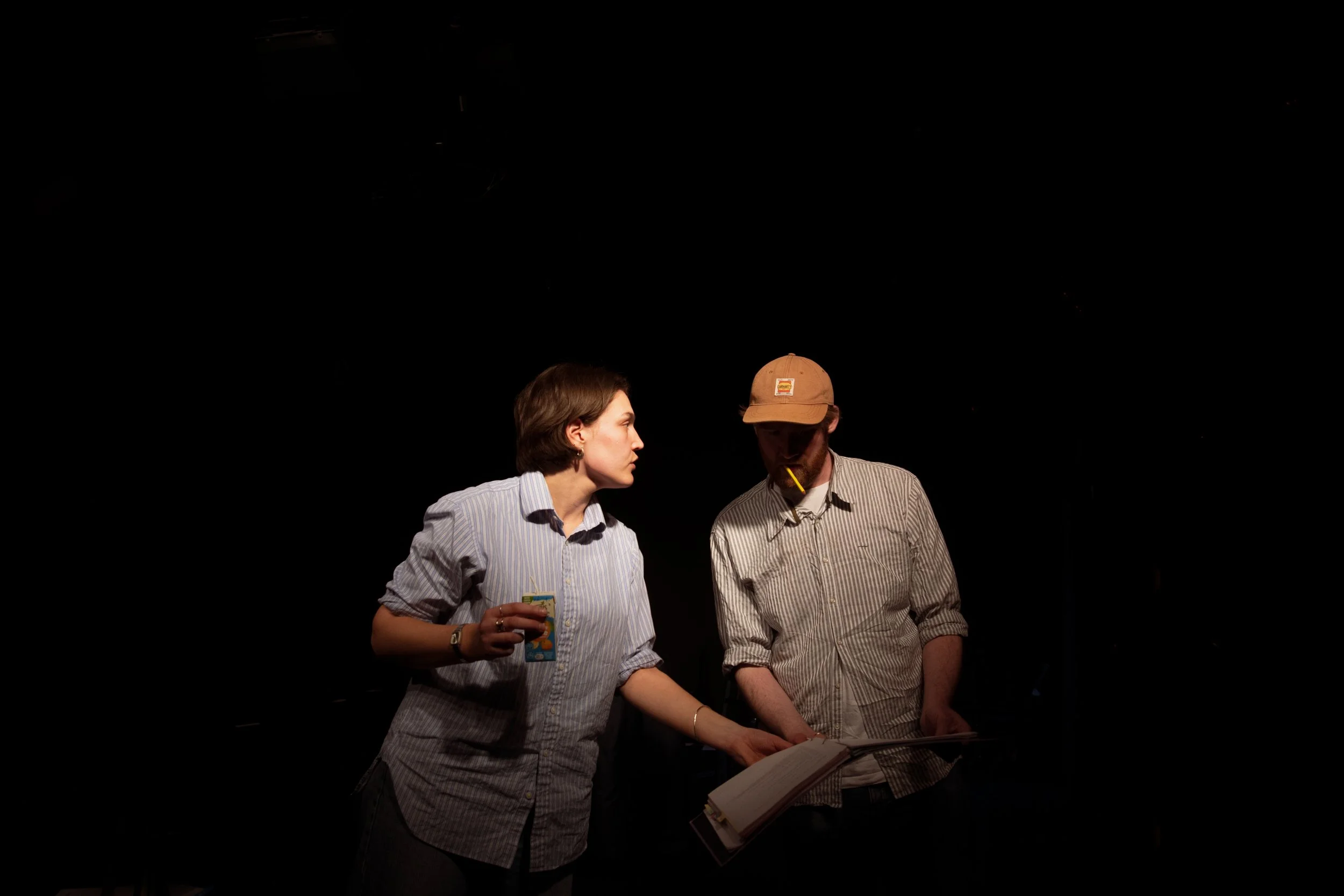 Two people, a woman and a man, appear to be engaged in a conversation or activity, with the woman holding a juice box and the man with a book and a visible pencil in his mouth, under spotlight against a dark background.