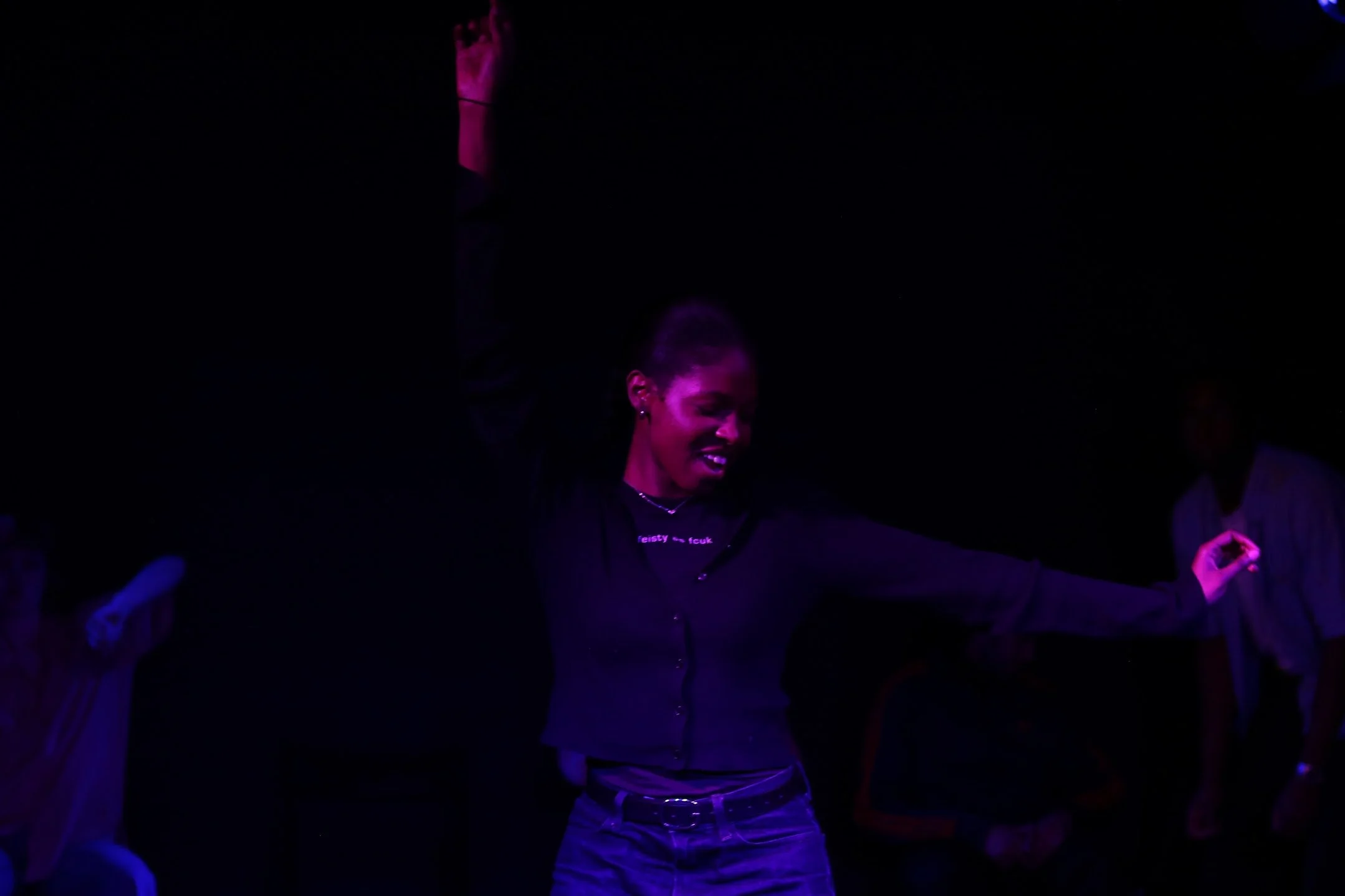 A person dancing with arms raised, smiling, in a dark setting illuminated with purple and blue lights.