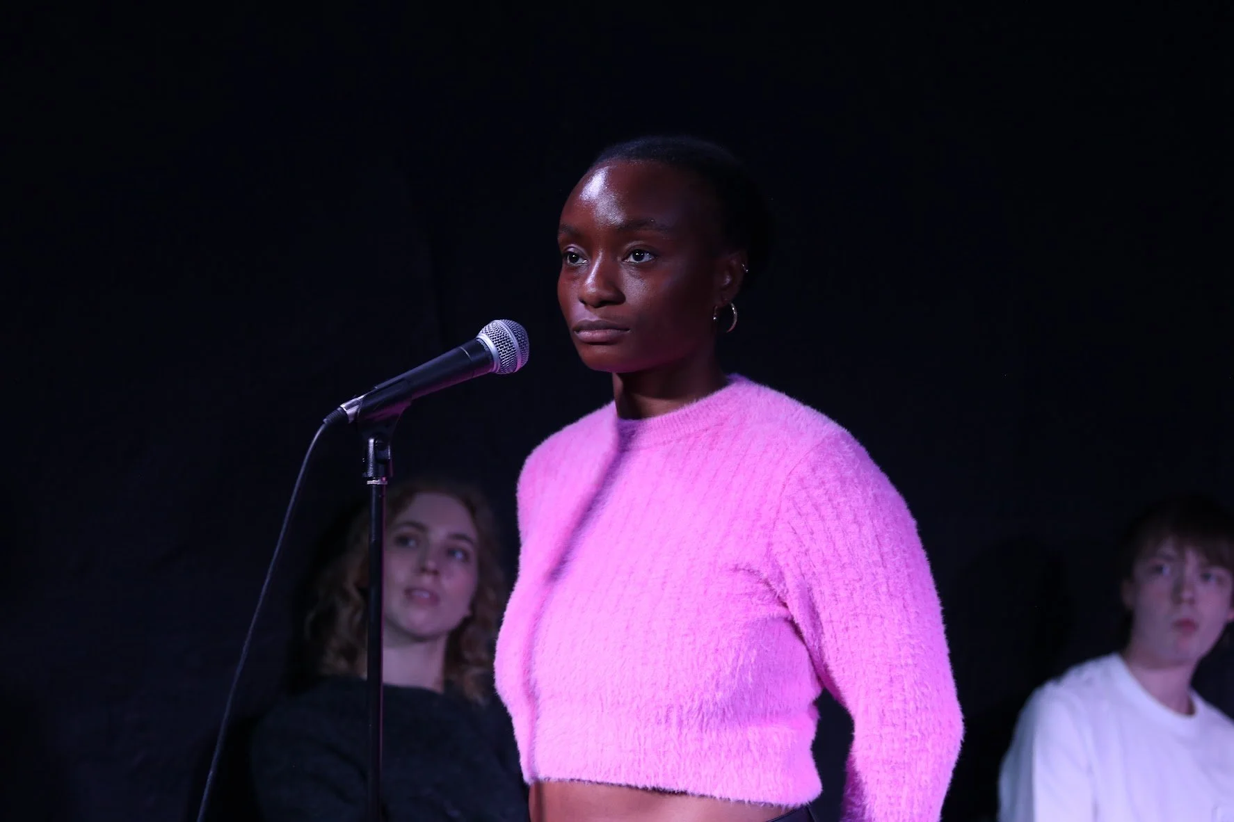 A young Black woman in a pink sweater stands in front of a microphone on a dark stage, with two young women behind her, including one with curly hair in a black top and another with straight hair in a white shirt.