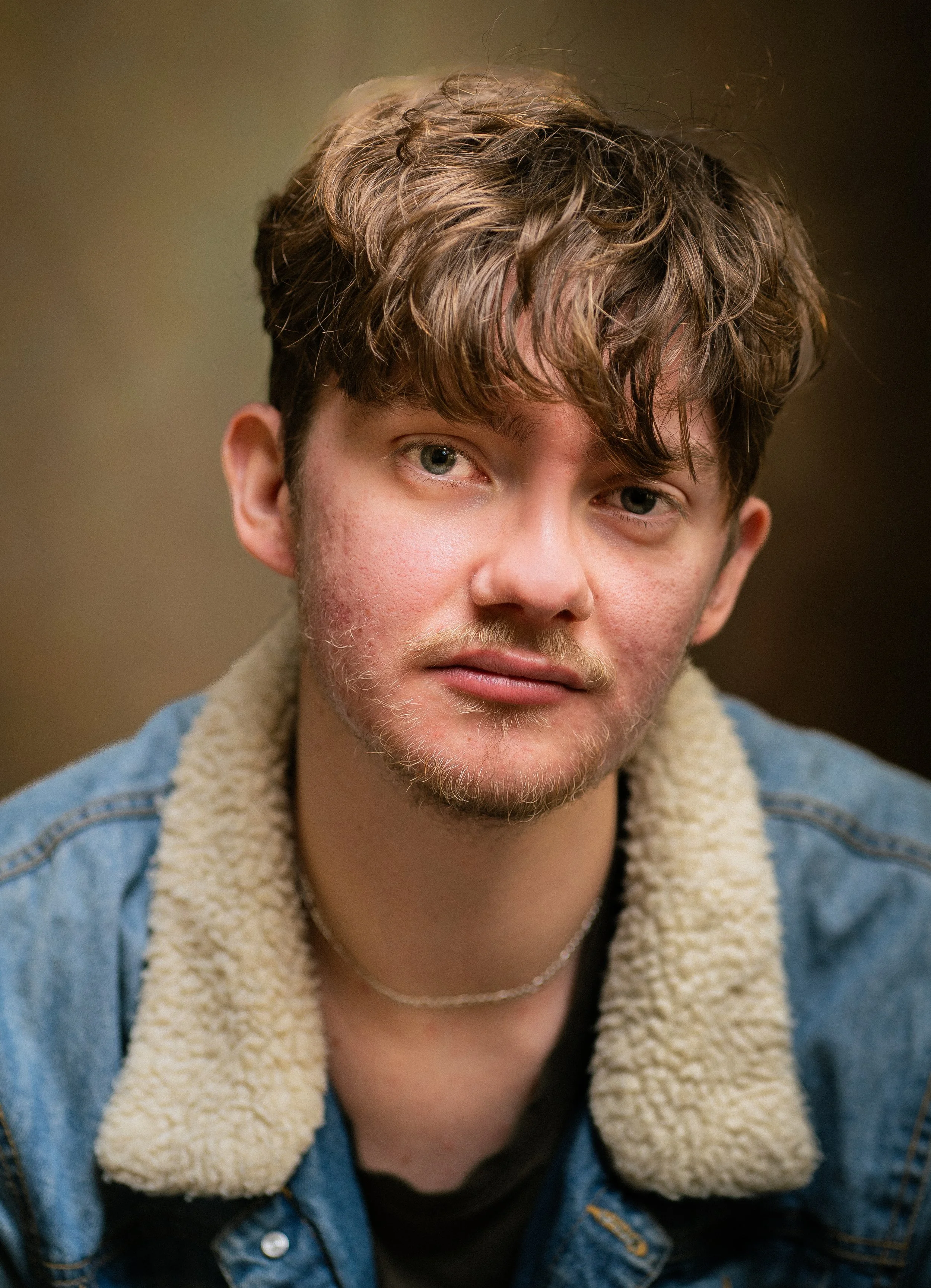 Close-up of a young man's face with light skin, green eyes, slight facial hair, and tousled brown hair, wearing a blue denim jacket with a sherpa collar and a black shirt underneath.