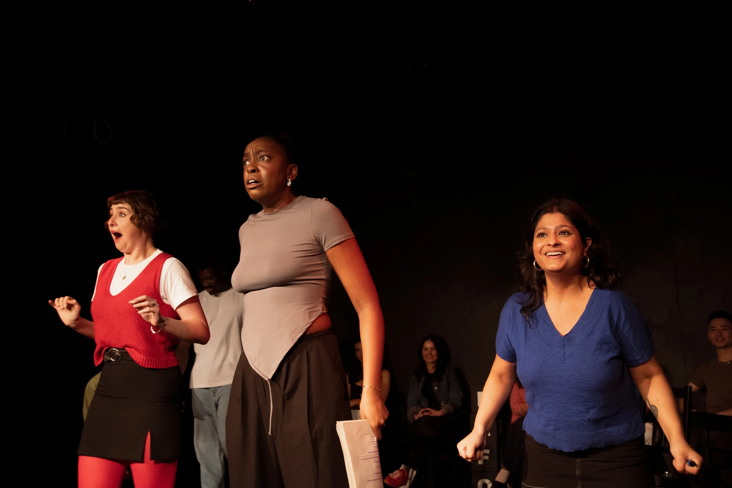Three women on stage, appearing surprised or excited, with an audience in the background.
