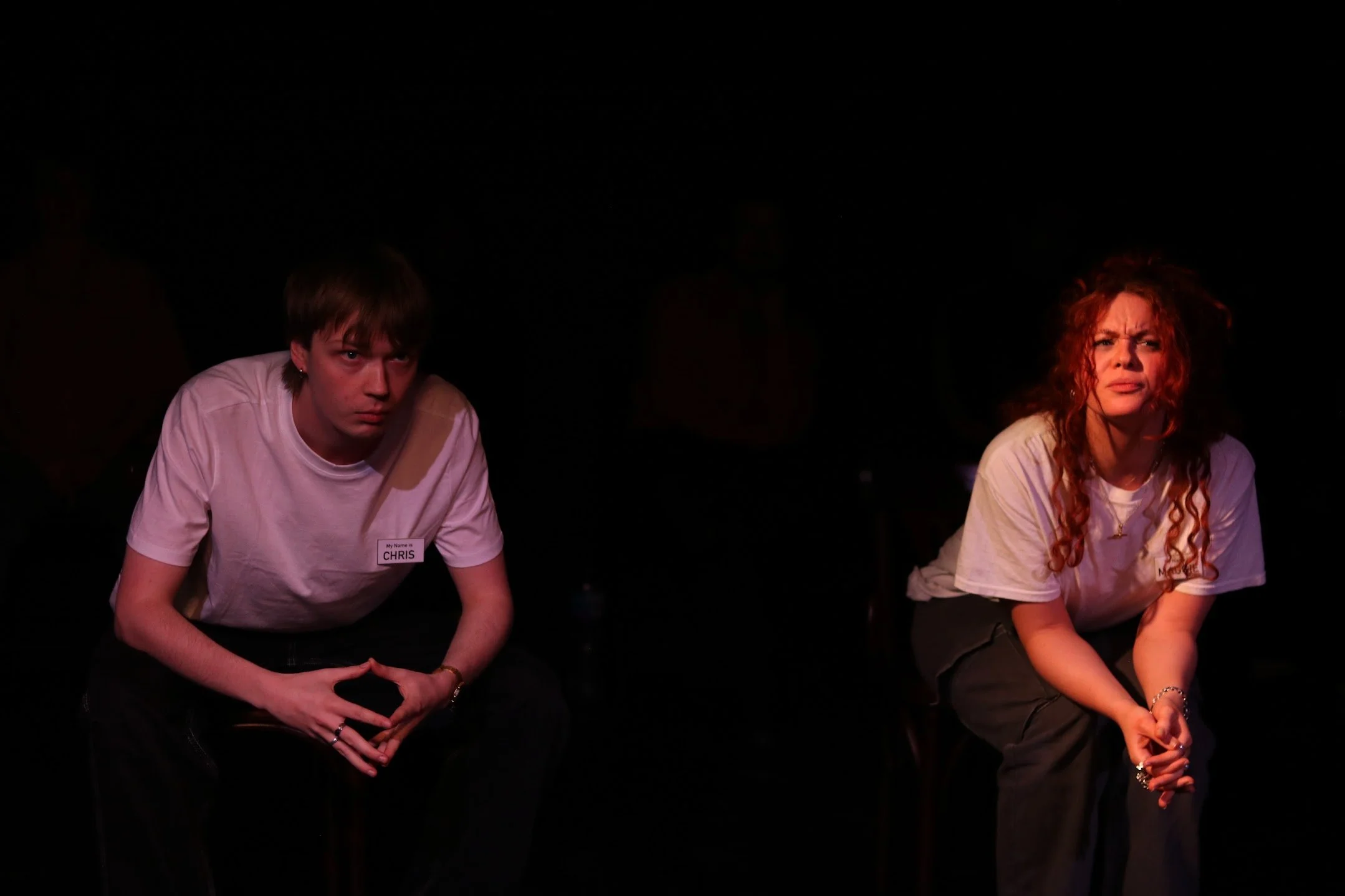 Two people sitting on a stage, appearing to be engaged or focused, with a dark background. The person on the left is a young male with short brown hair, wearing a white t-shirt and a name tag that reads 'Chris.' The person on the right is a young female with curly red hair, wearing a white t-shirt.