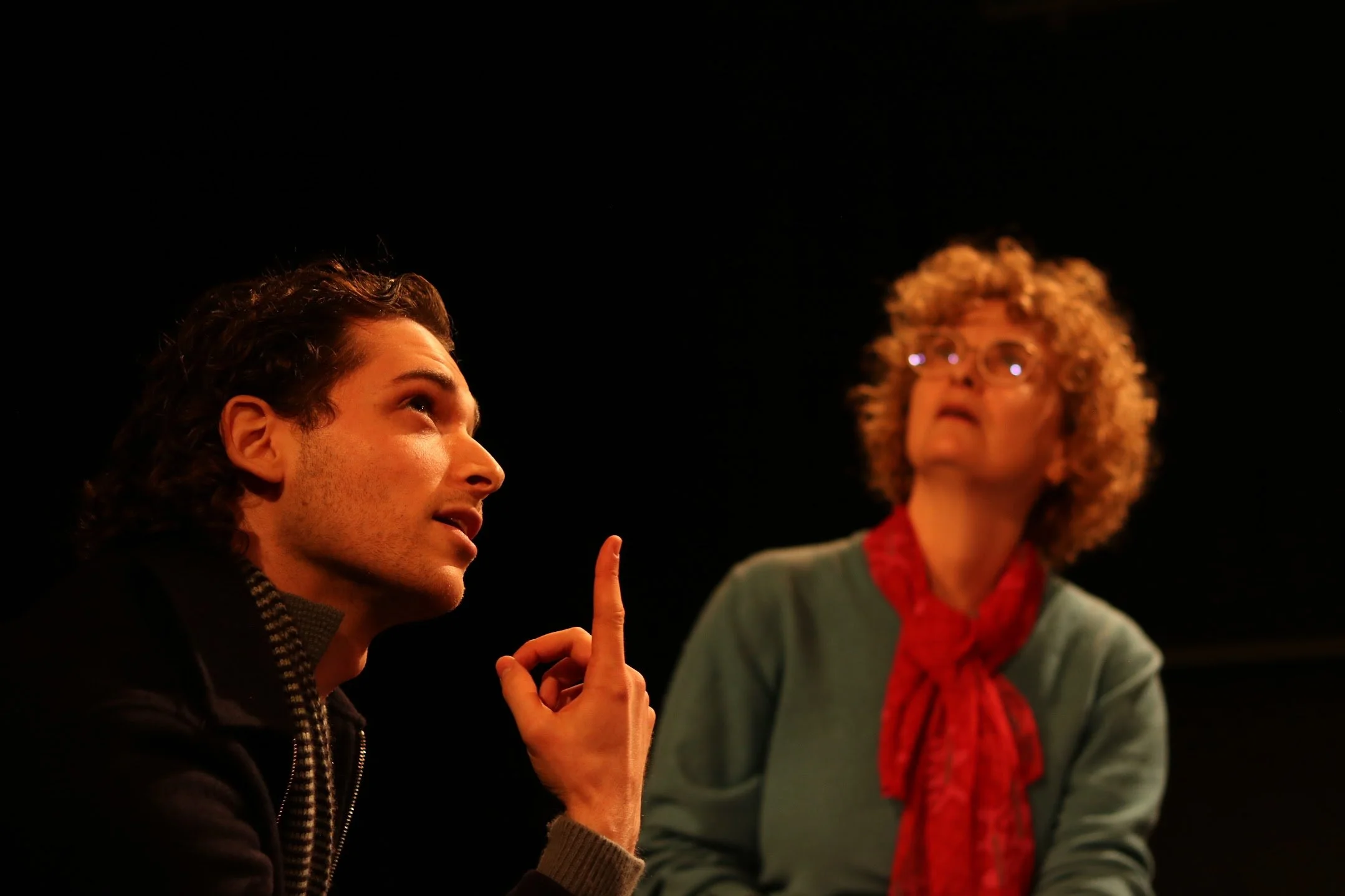 A young man with curly brown hair speaking and gesturing with his right hand, while a woman with curly blonde hair and glasses listens, in a dark setting.