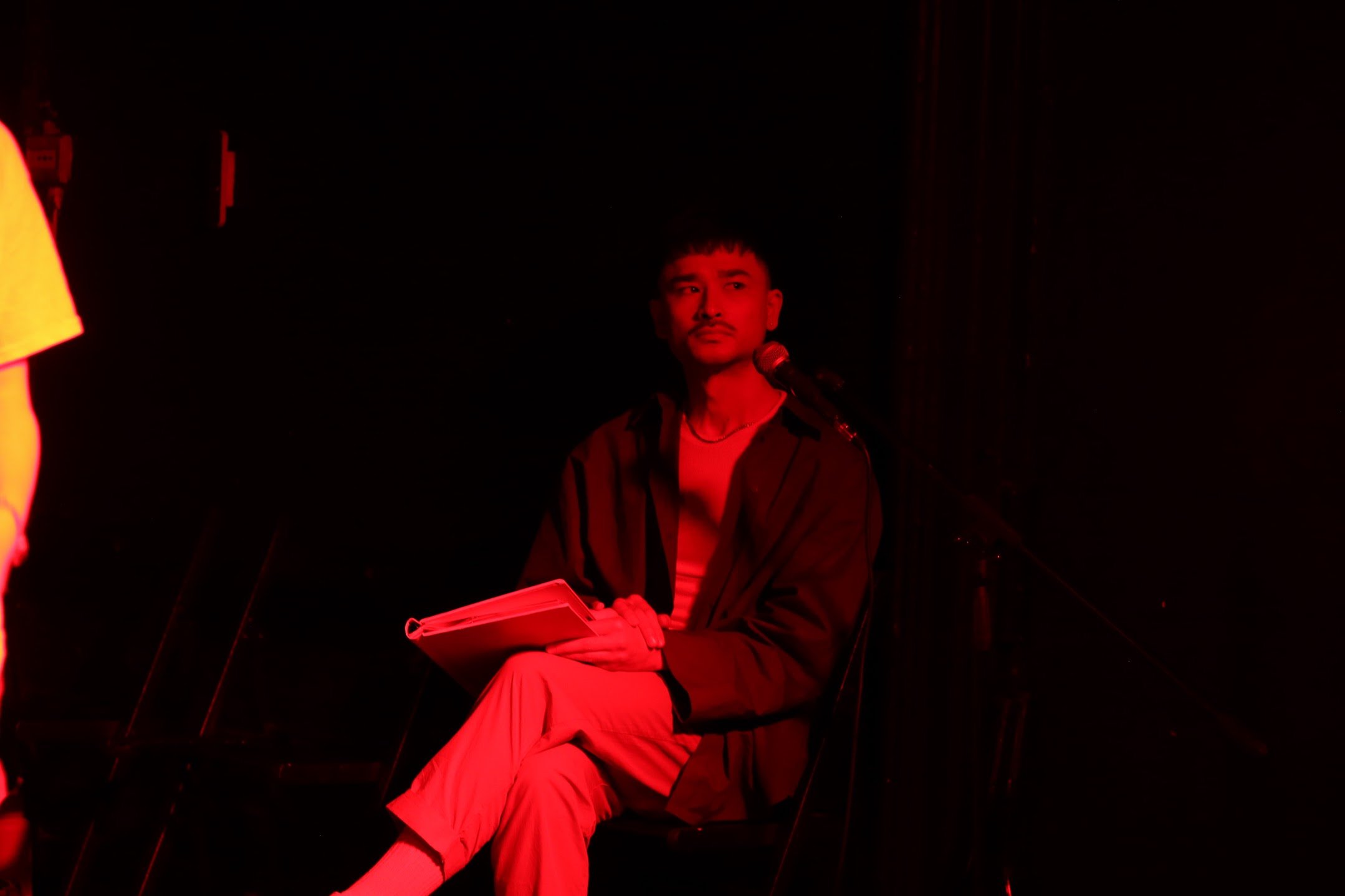 A person sitting with their legs crossed, holding a notebook, in a dimly lit room with red lighting.