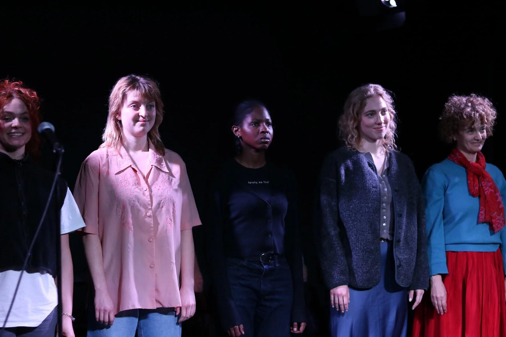 Five women standing in a line on a stage, facing forward, with a dark background. They are dressed casually, and the woman on the left is speaking into a microphone.