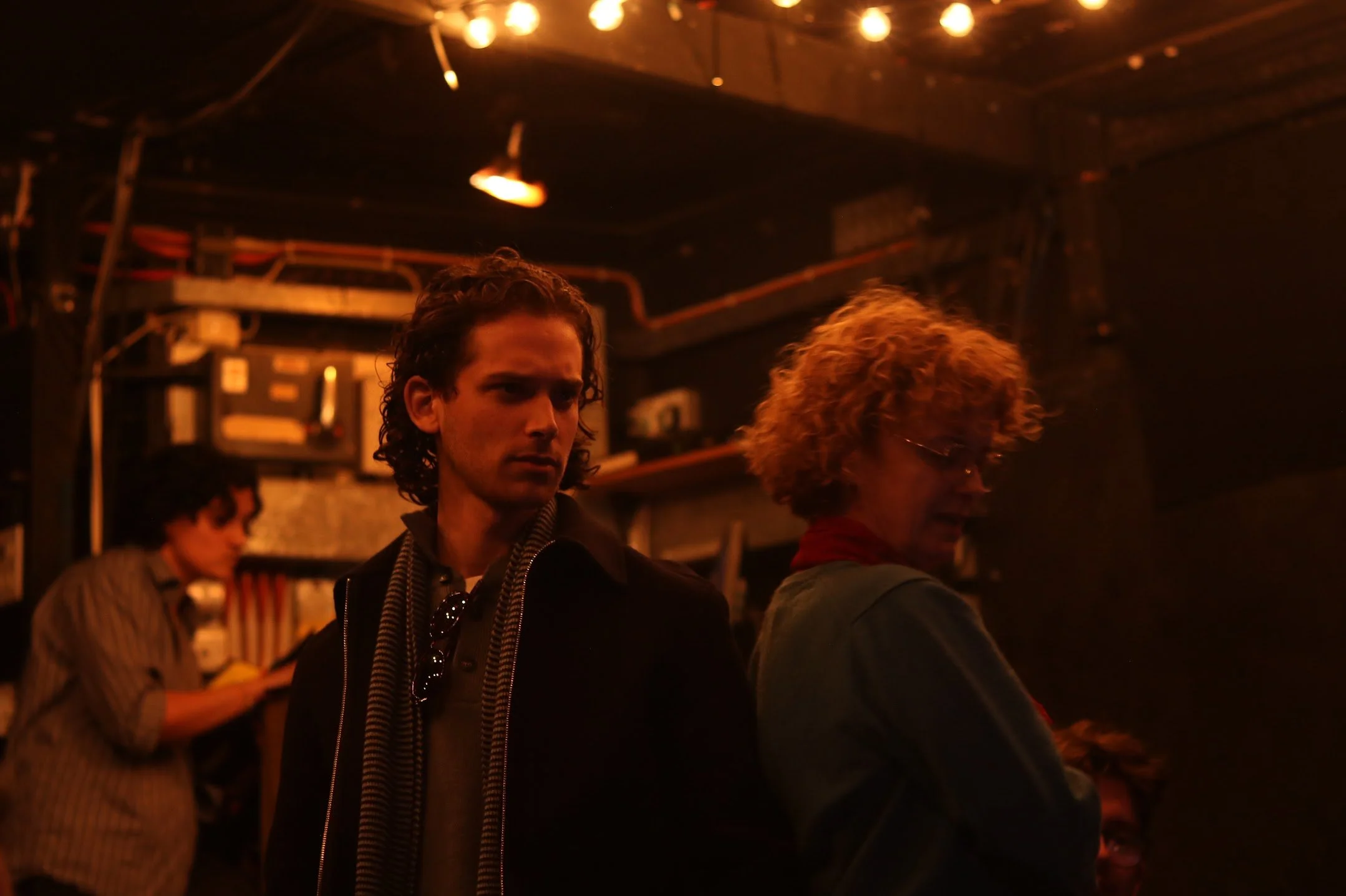 Three people are in a dimly lit bar. One man with curly hair and a dark jacket is looking slightly to the side, and a woman with short curly hair and glasses is standing next to him. In the background, another person is focused on something, possibly
