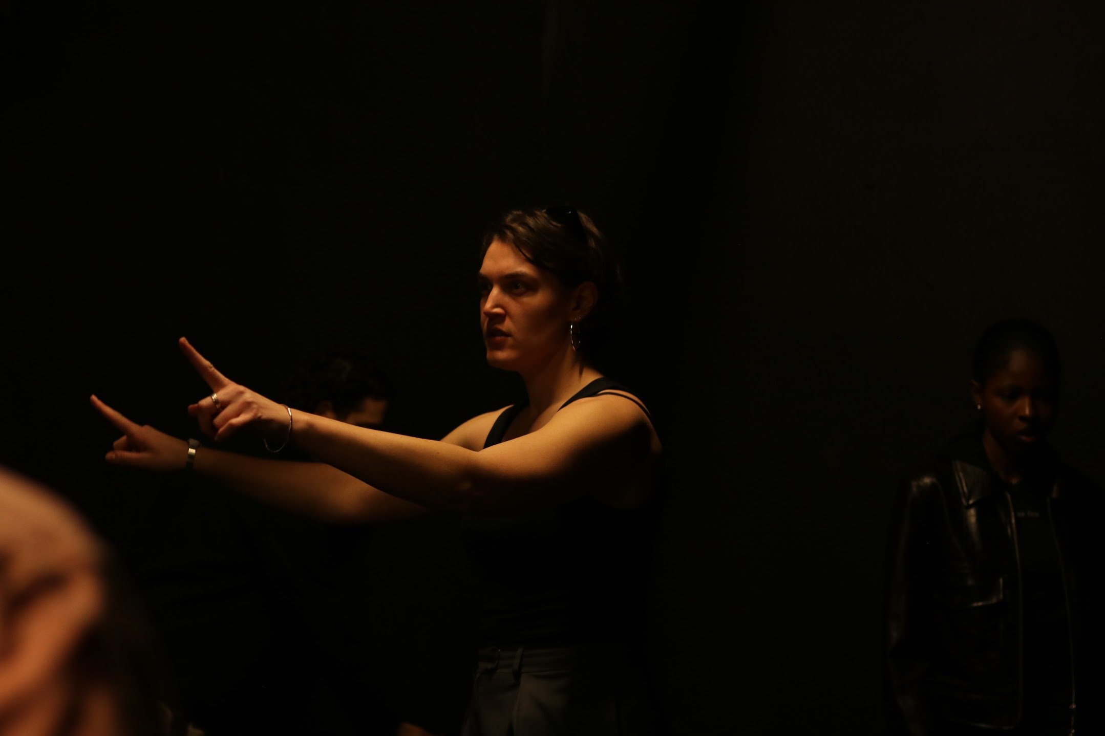 A woman with short dark hair and earrings is pointing to the left in a dark room, with a few other people partially visible around her.