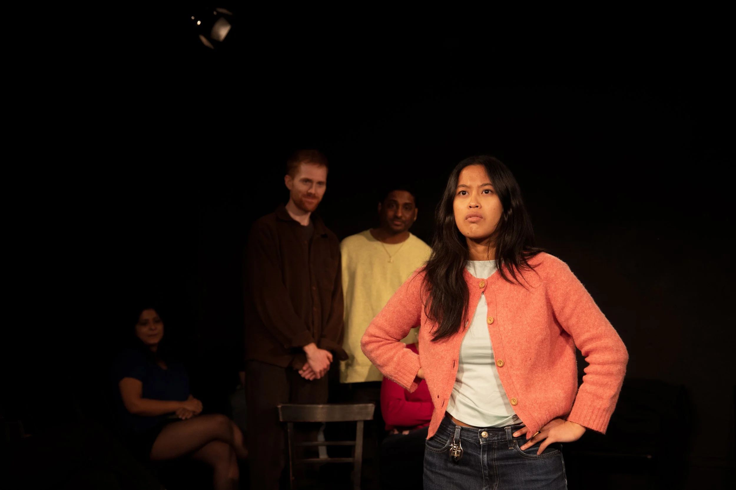 A young woman with long dark hair standing with her hand on her hip in front of a dark background, wearing a light-colored shirt and pink cardigan. Behind her, three people are visible but slightly out of focus: a man with short hair and a beard wear