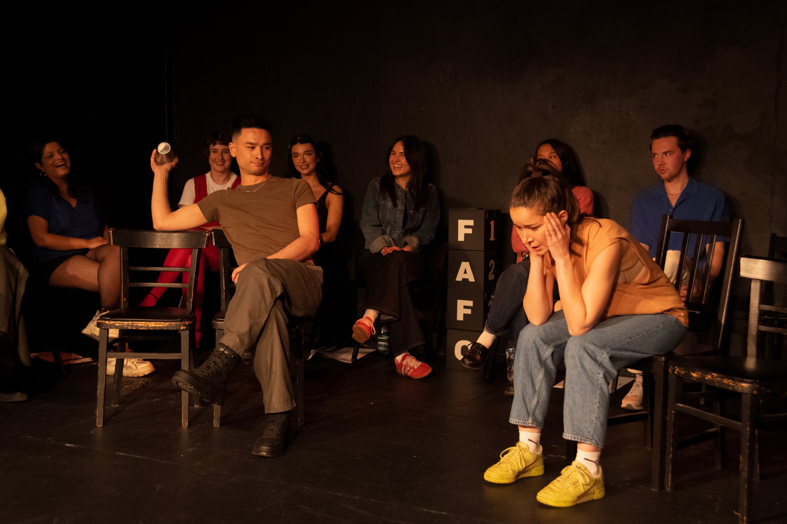 A group of diverse young adults sitting and standing on a stage with a dark background, some laughing and smiling, one looking unimpressed, and one looking distressed, with theater blocks spelling 'FACE' on the floor.