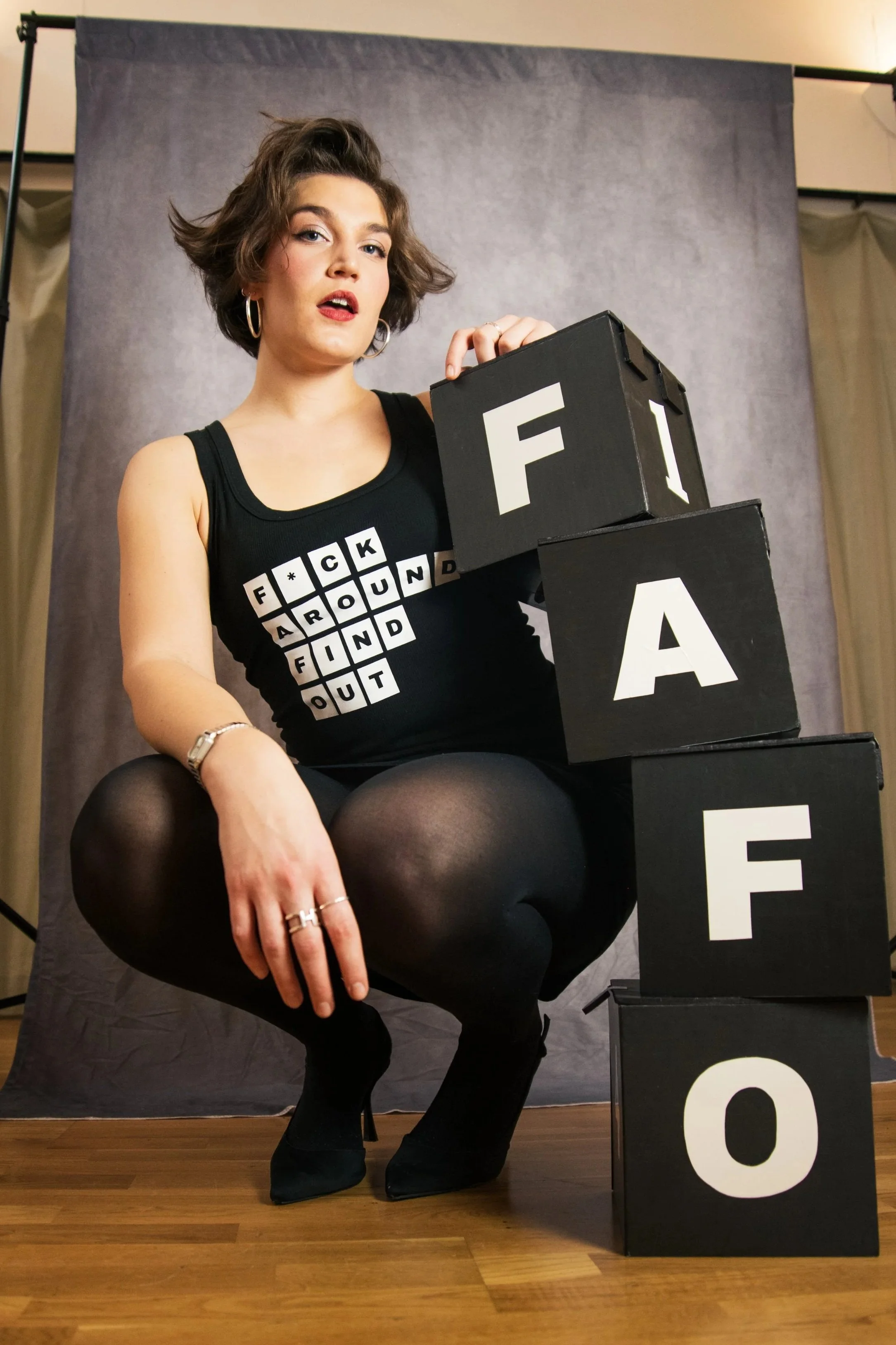 A woman with short, wavy brown hair and hoop earrings is sitting on a stack of black boxes with letters on them. She is wearing a black tank top with a crossword puzzle design, black tights, and black shoes. The background is a grey fabric backdrop.