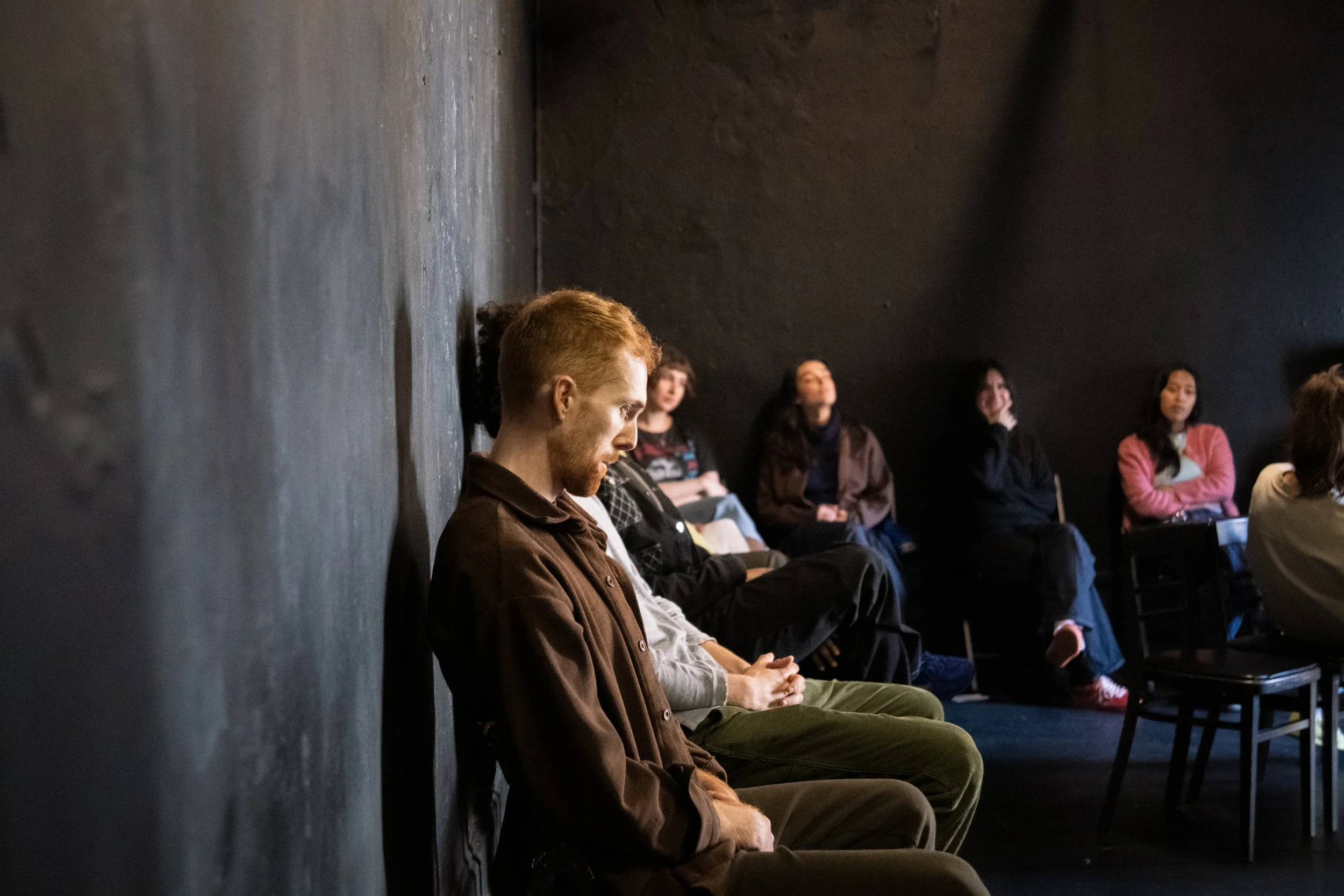 People sitting with eyes closed or looking down against a dark wall in a room with black walls.