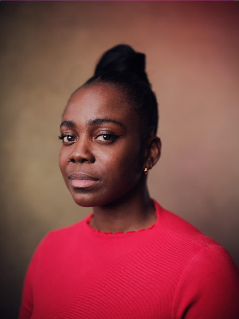 Portrait of a Black woman with neatly styled hair in a bun, wearing a red top, against a blurred brownish background.