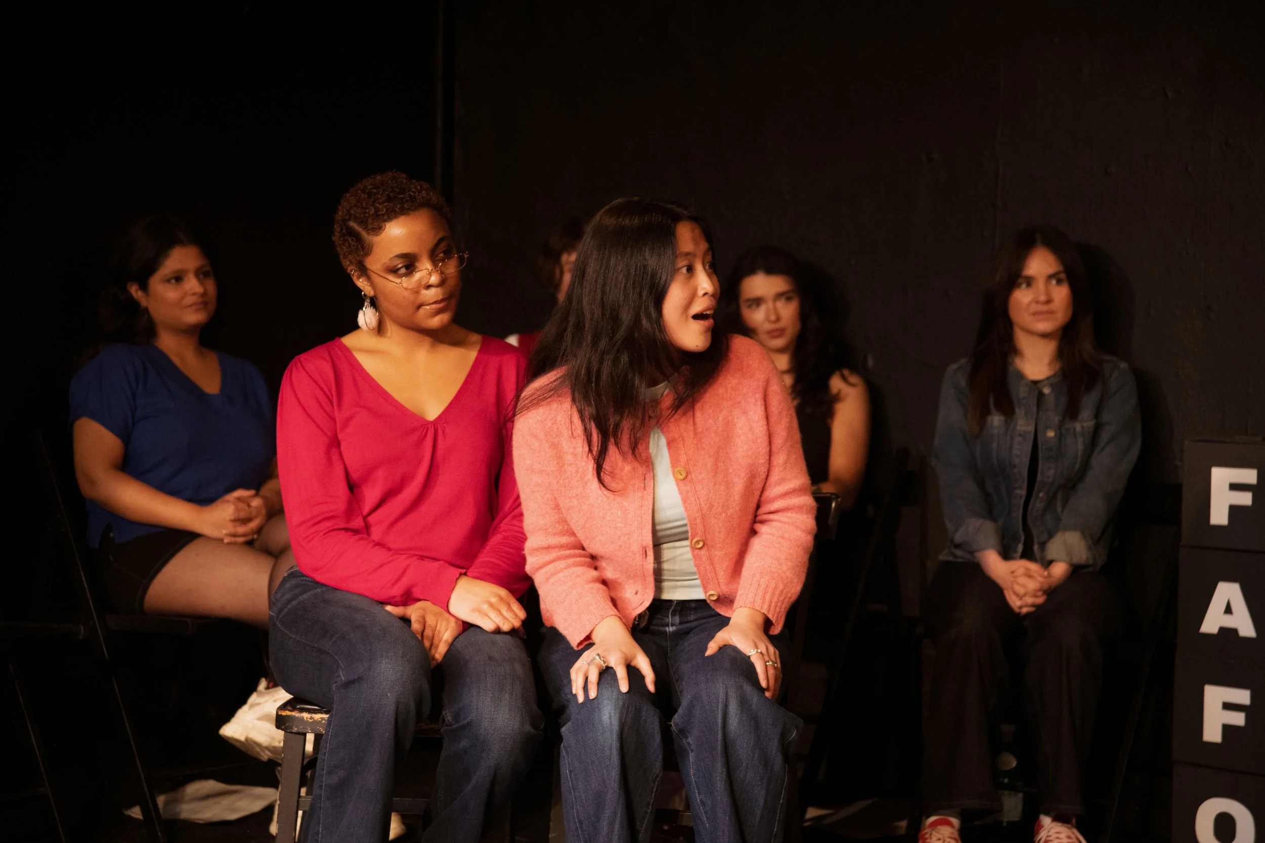 Six women sitting and watching an event against a black background.