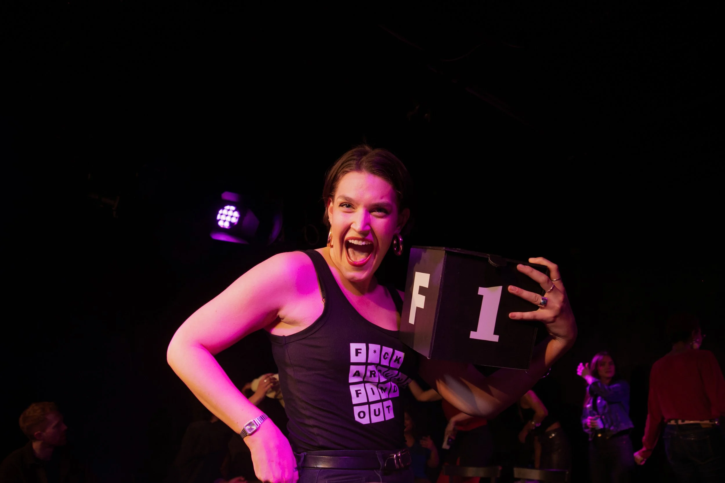 A woman with short brown hair, earrings, and a purple-lit face, smiling and showing excitement, holding a chalkboard sign with 'F 1' written on it at a dark indoor event.