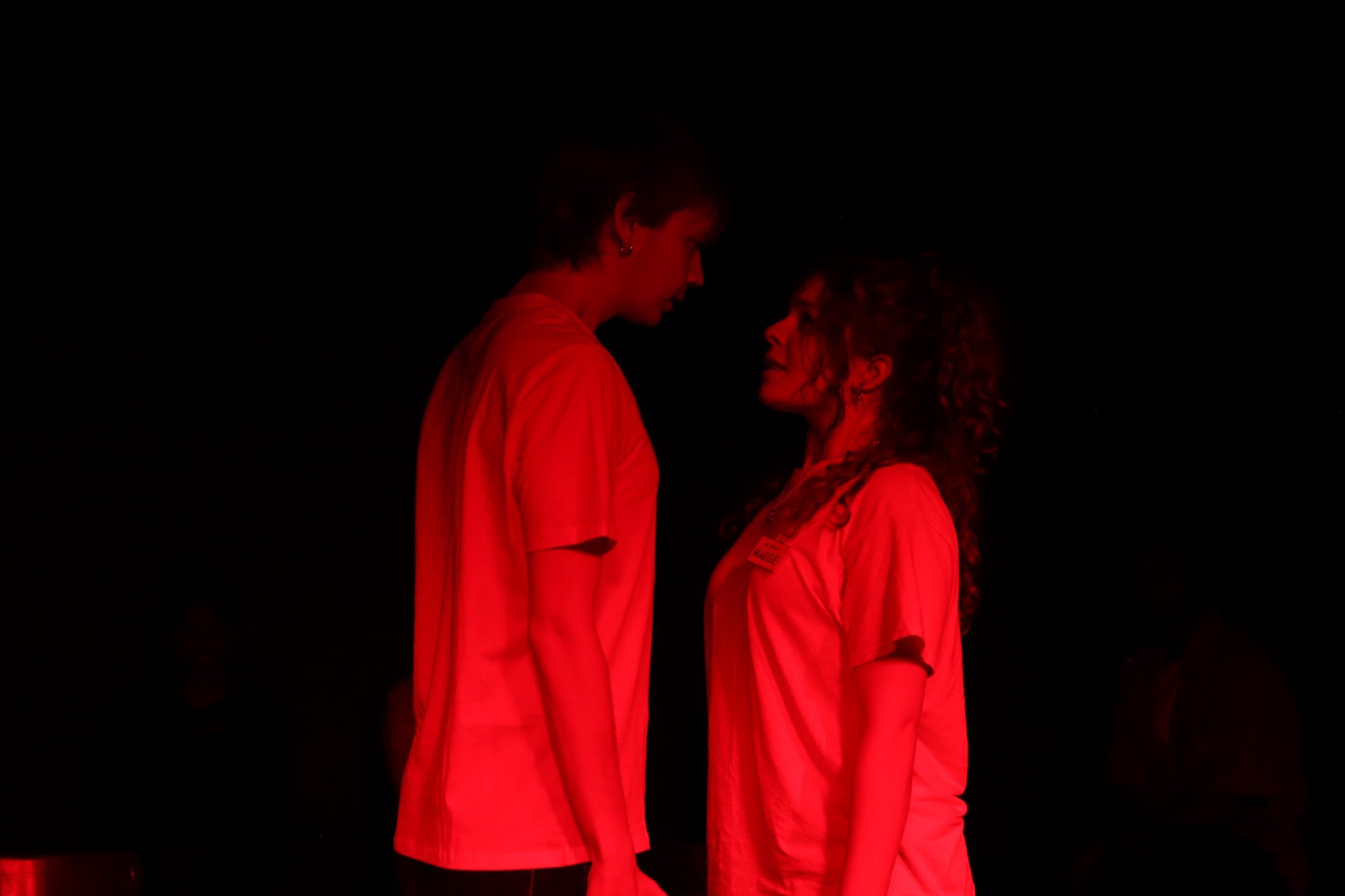 Two women face each other closely in red lighting, with one wearing earrings and a name badge.