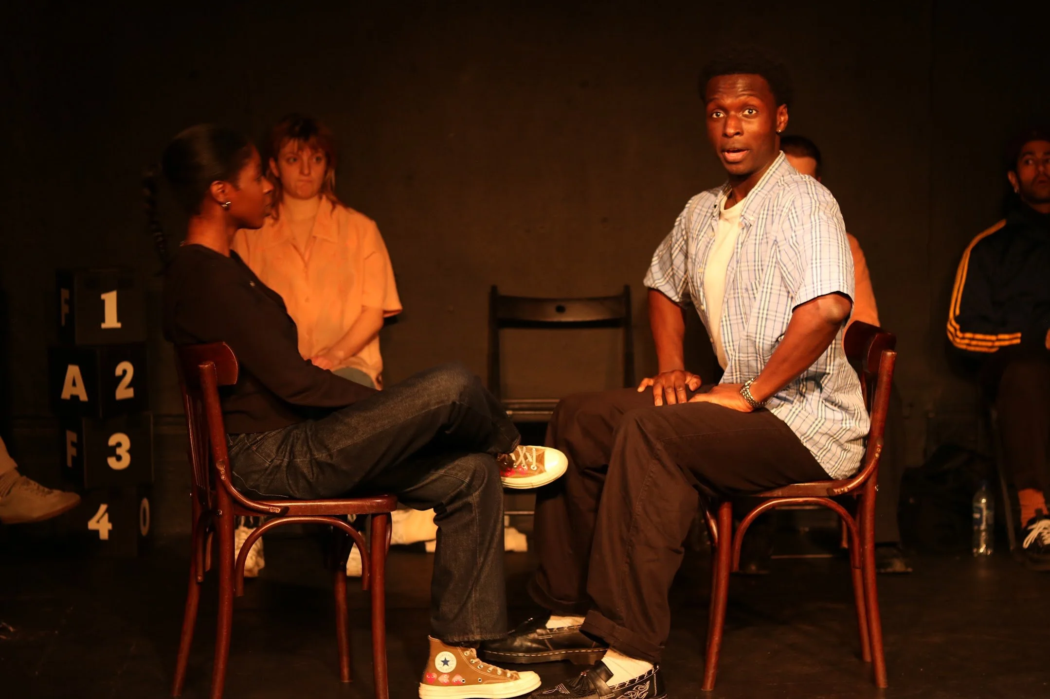 A stage play scene with five actors, one seated and four standing in a dark room. The seated actor has their legs crossed, wearing jeans and Converse sneakers with colorful patches. The standing actor to the right is wearing brown pants and a plaid s