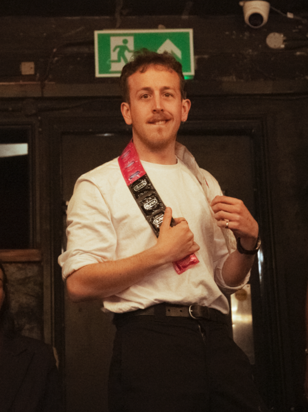 A man in a white shirt and black pants, holding a multi-pack of snacks over his shoulder, standing in front of a dark door in an indoor setting with emergency exit sign above him.