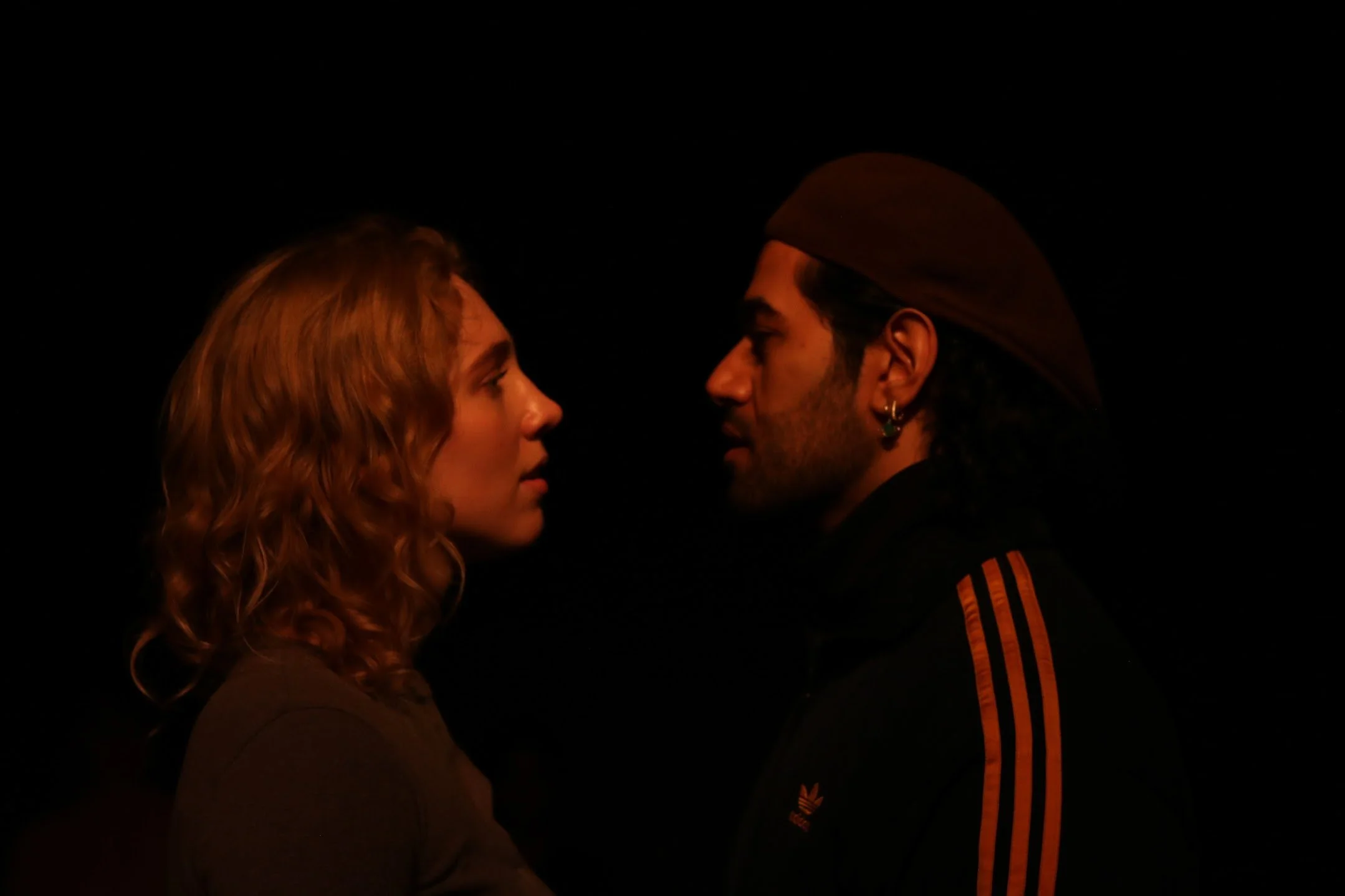 A woman and a man face each other in a dark setting, close up, with minimal lighting highlighting their faces.