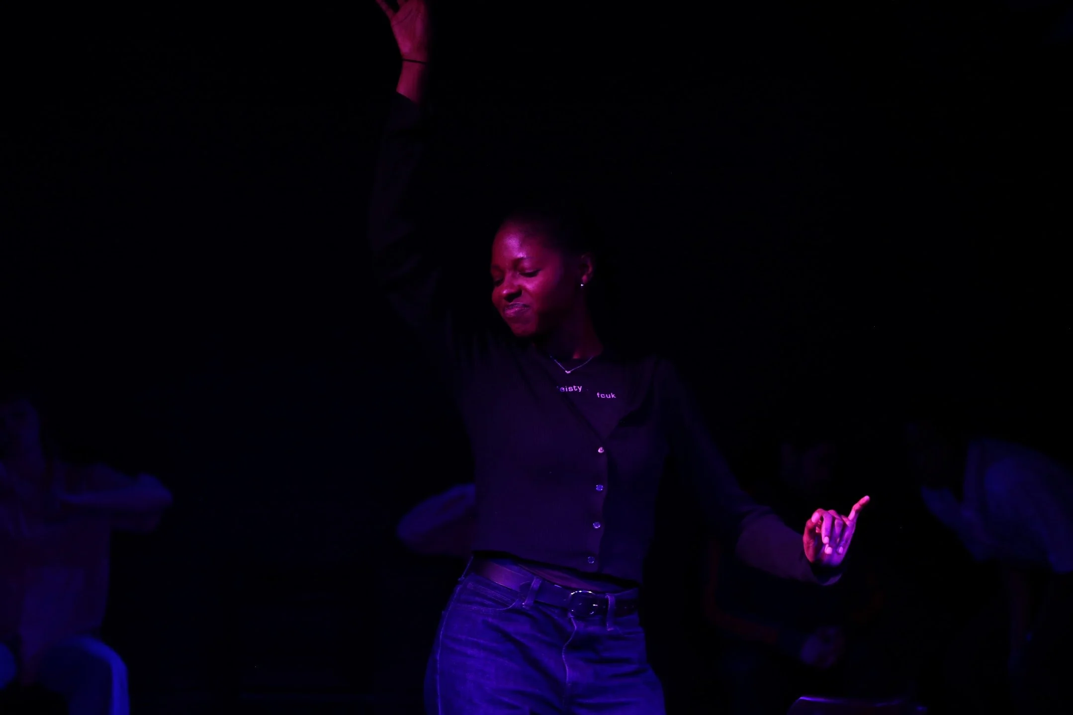 A woman with closed eyes, smiling, wearing a black shirt with white text, dancing in a dark setting with purple lighting.