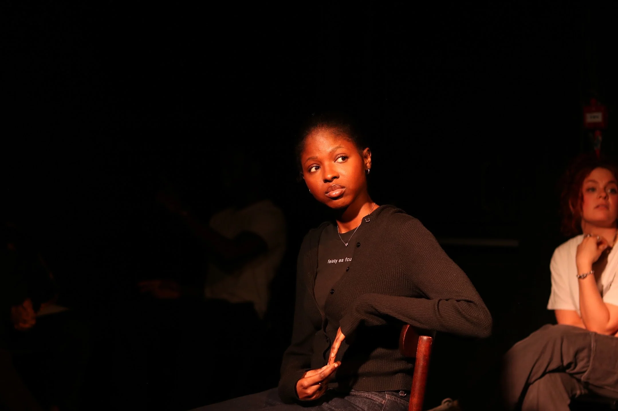 A young woman with dark skin and wearing a black top sitting in a dimly lit environment, looking to her left. She has earrings and a necklace, and her hair is styled short and curly. There are two women visible in the background, one on the right with red hair and another partially visible on the left, all in a dark setting with minimal lighting.