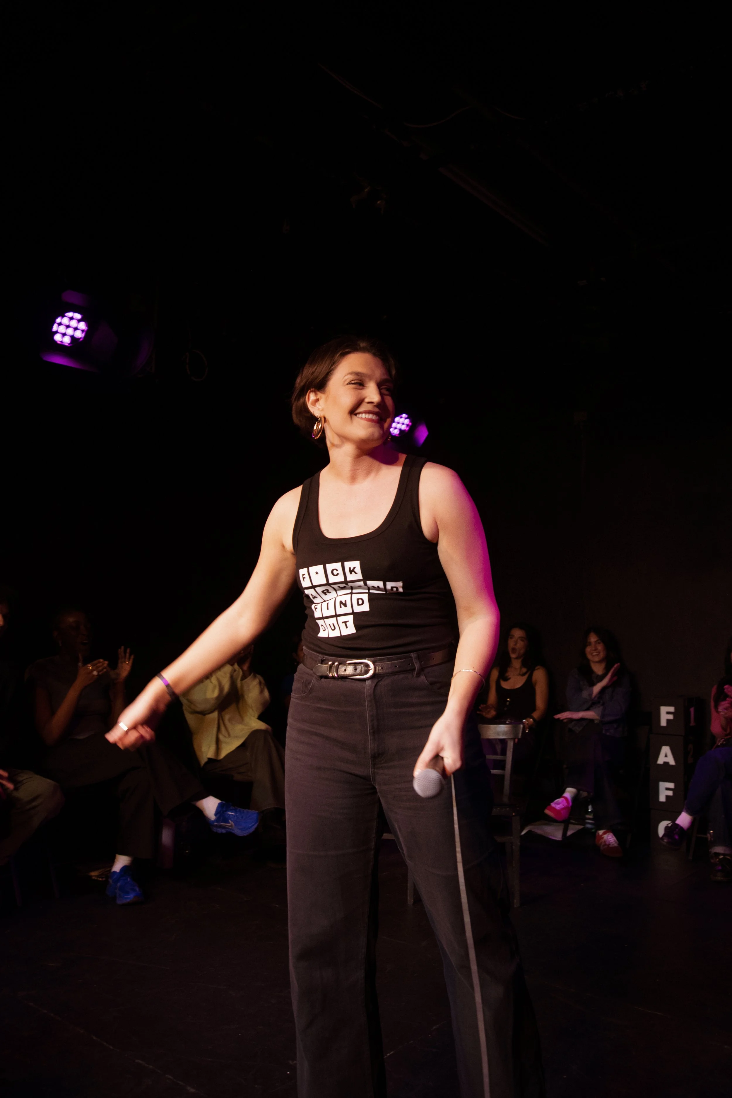 A woman with short brown hair, wearing hoop earrings, a black tank top with a provocative phrase, and black pants, holding a microphone and smiling, standing on a stage with purple stage lights, surrounded by an audience.