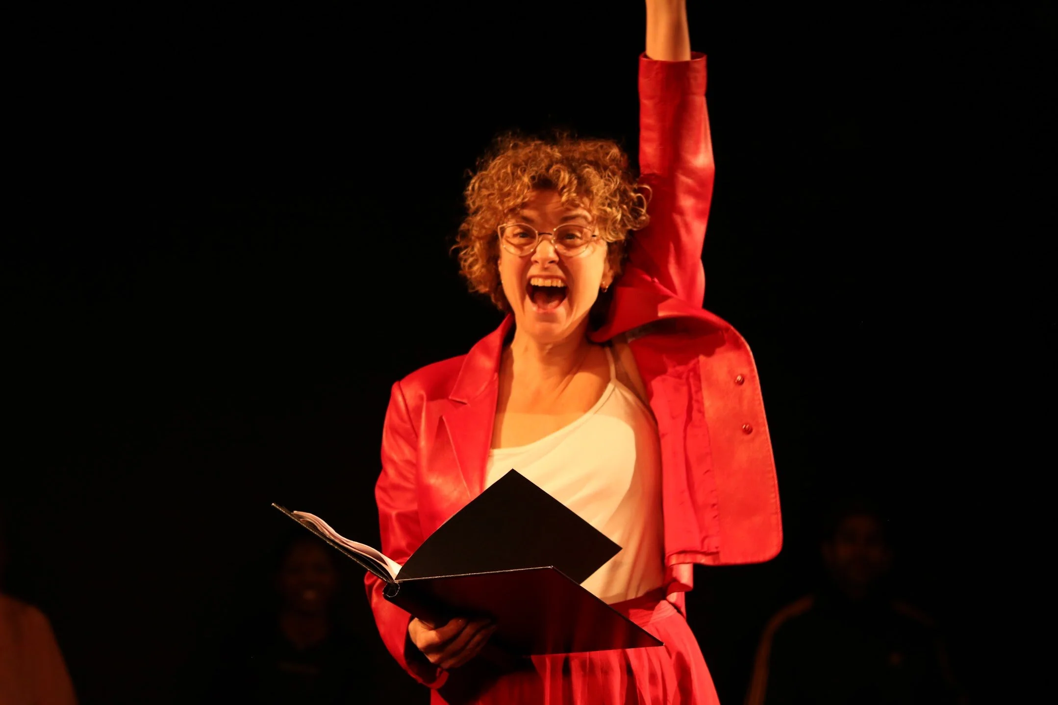 A woman with curly hair and glasses singing passionately while holding a black folder, wearing a red jacket in a dark setting.