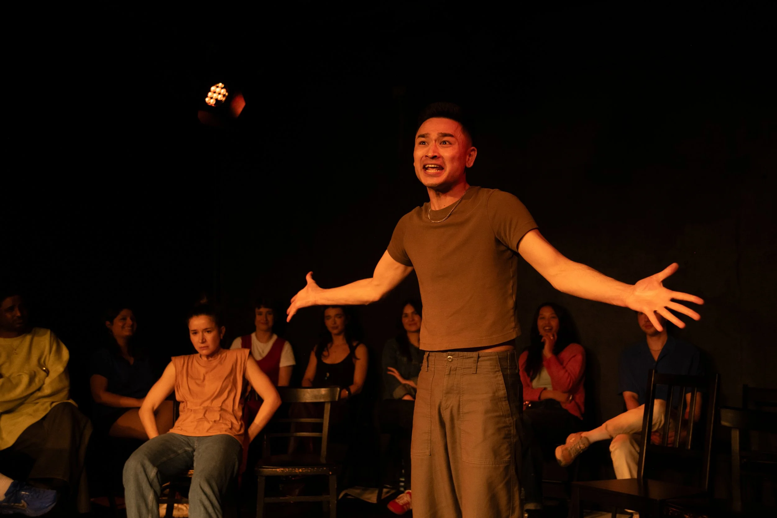A man with short dark hair and wearing a brown t-shirt and pants is performing on stage with his arms outstretched. He is surrounded by an audience of diverse people seated and watching him. The background is dark.