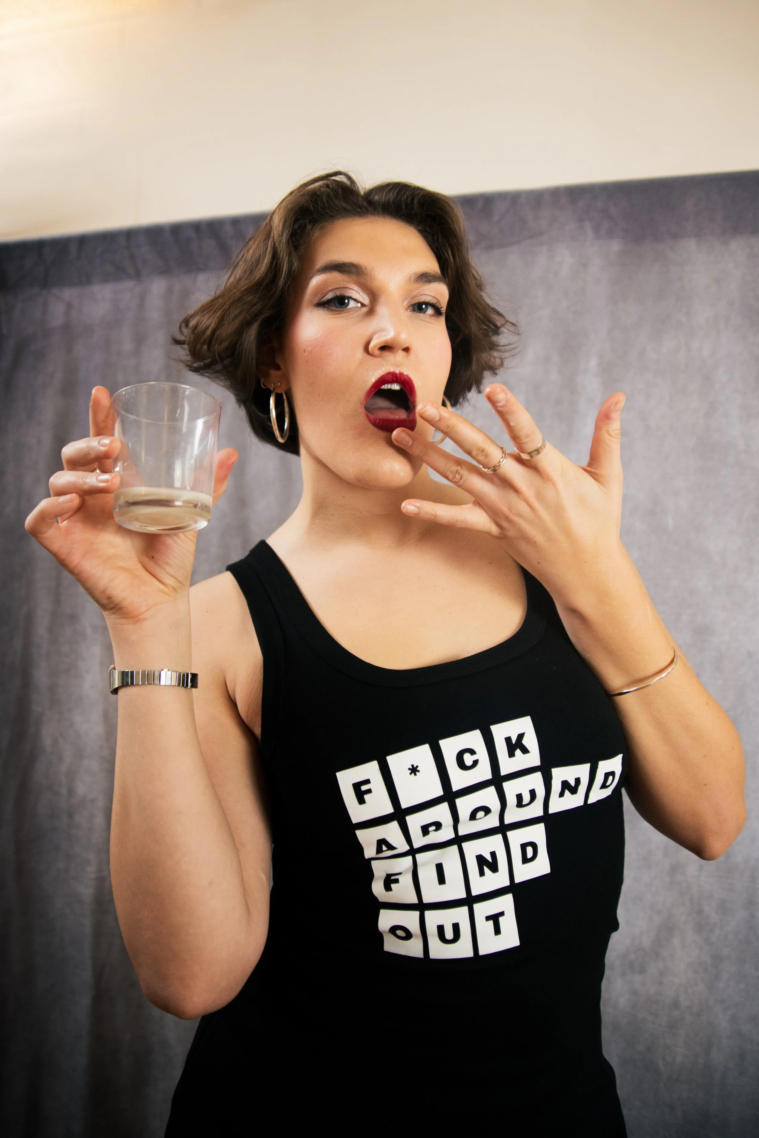 A woman with short brown hair, wearing a black tank top with a Scrabble-inspired design, is holding a glass with a clear drink in her right hand and touching her lips with her left hand. The background is a neutral gray curtain.