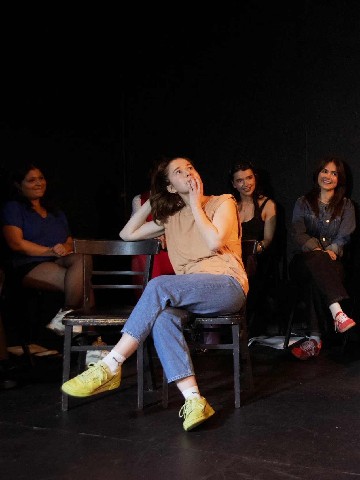 Group of women sitting on chairs against a black background, one woman in the foreground wearing a beige top and yellow sneakers, looking surprised or curious.