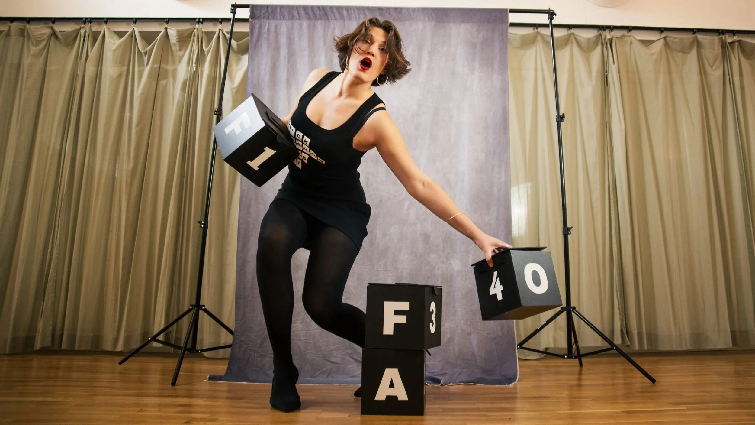 A woman with short brown hair wearing black clothes and pose with several large black blocks with white letters and numbers, standing on a wooden floor with beige curtains in the background.