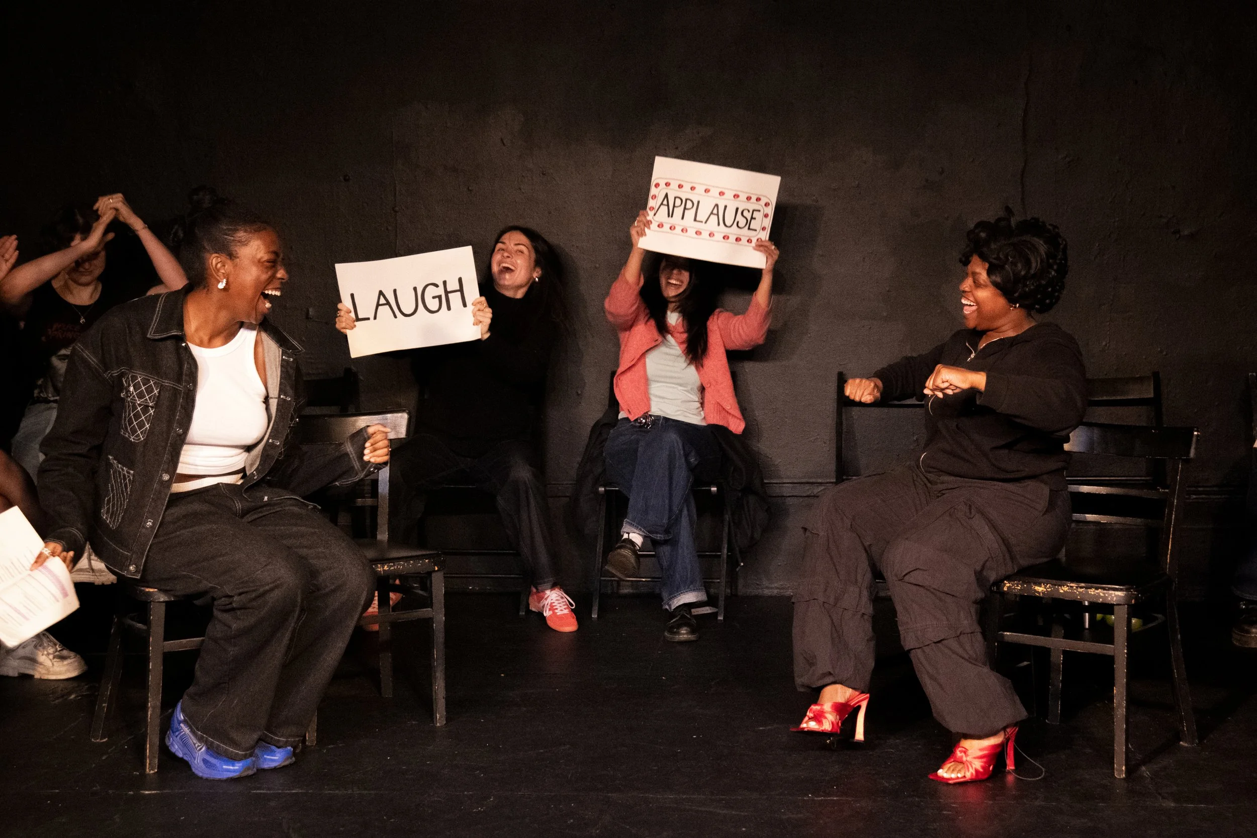Group of women laughing and smiling during a lively social gathering, one holding a sign that says 'LAUGH,' and another holding a sign that says 'APPLAUSE,' in a dark room with black walls.