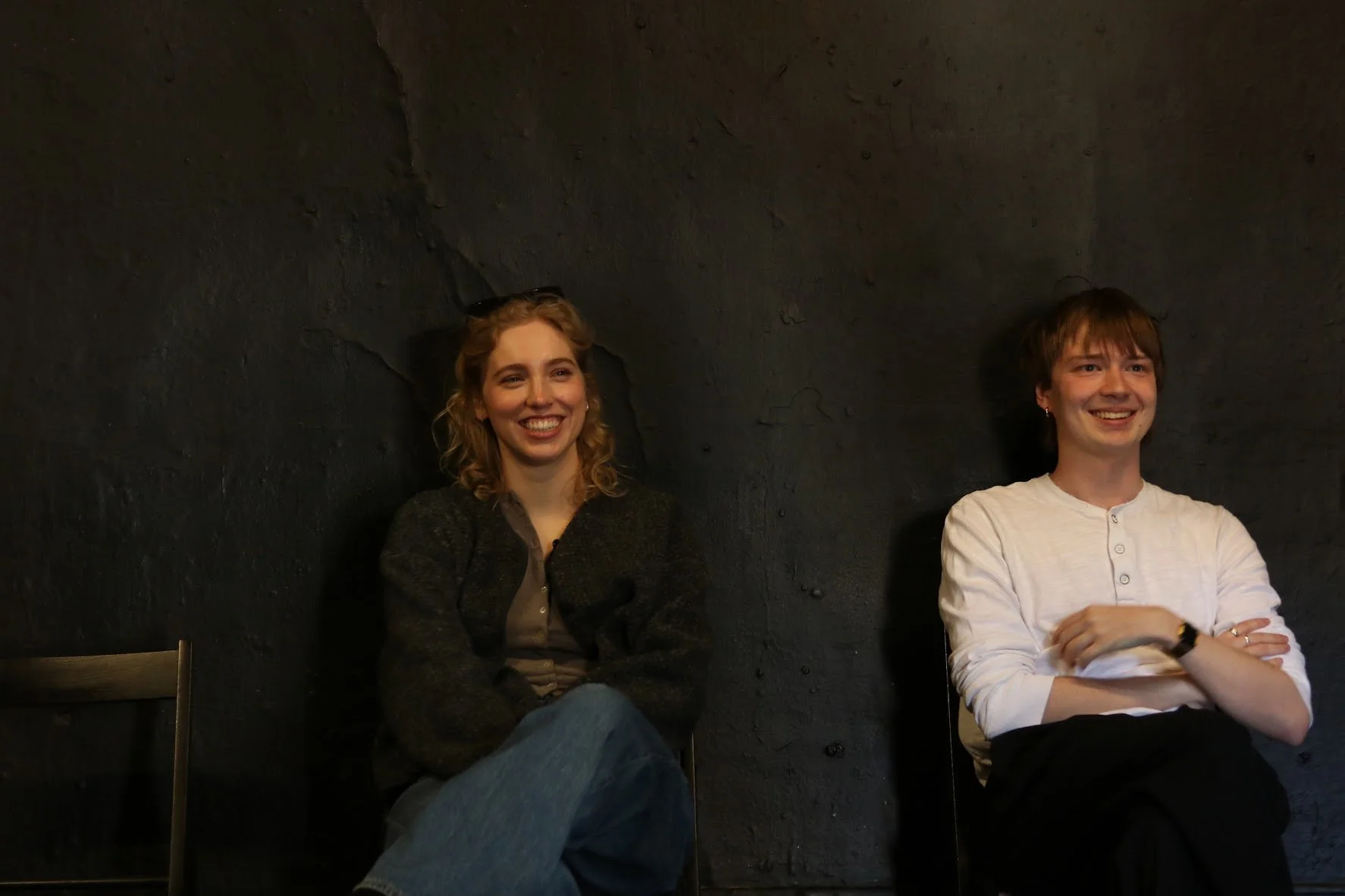 Two smiling young women sitting against a black textured wall, one with blonde hair wearing a dark sweater, and the other with short brown hair wearing a white shirt, both appearing relaxed and happy in an indoor setting.