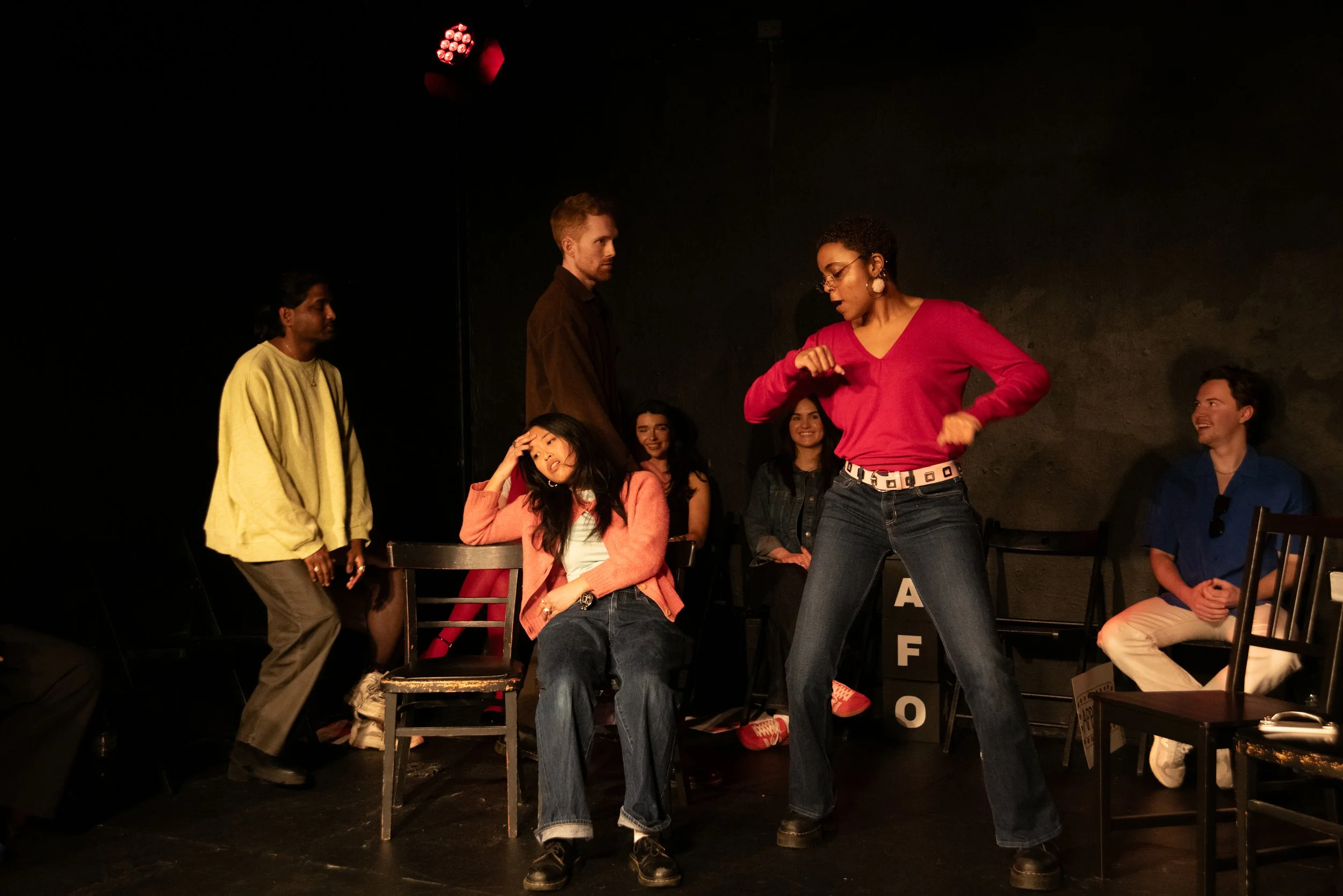 A group of actors and actresses on a theater stage, with some seated and some standing. One woman in a red top and jeans appears to be acting or dancing, while others observe and react. The background is black.