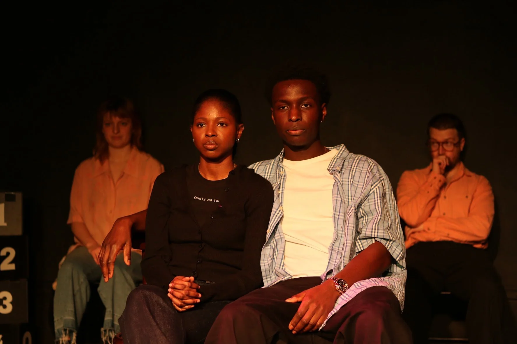 Four people seated on a stage in a black background, with two women in the back and a man and a woman in the front, appearing serious or contemplative.