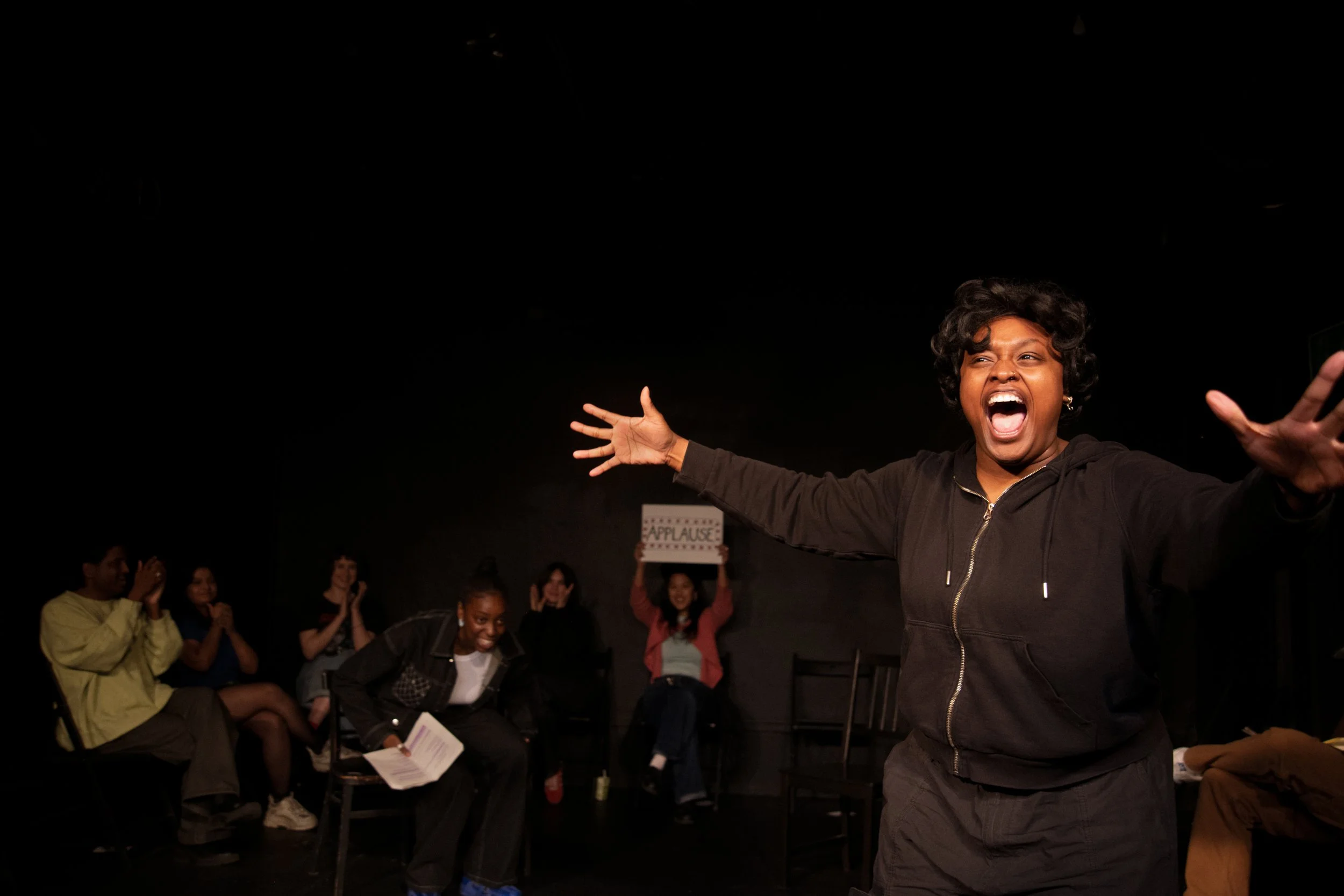 A woman with curly hair and a black zip-up jacket passionately performs on stage with arms broad open, a joyful expression, and an audience clapping in the background. The audience members are smiling and clapping, with some holding papers or signs.