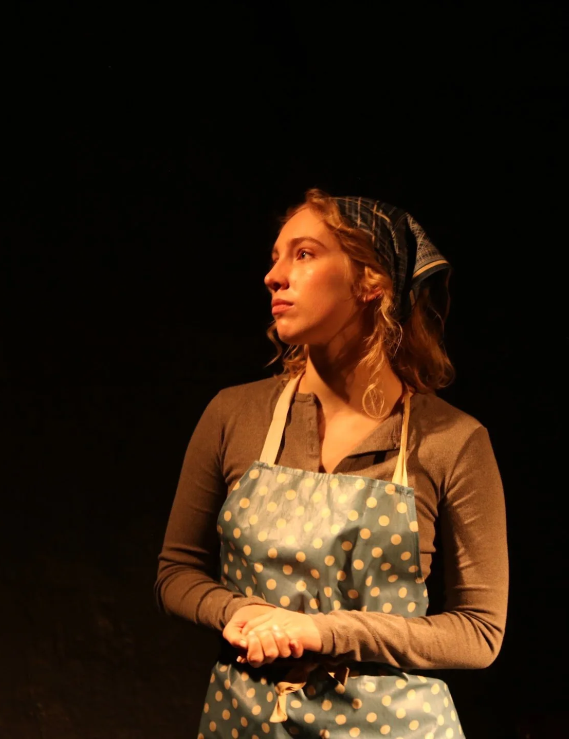 A woman wearing a polka dot apron and headscarf, standing against a black background, looking to the side with a serious expression.