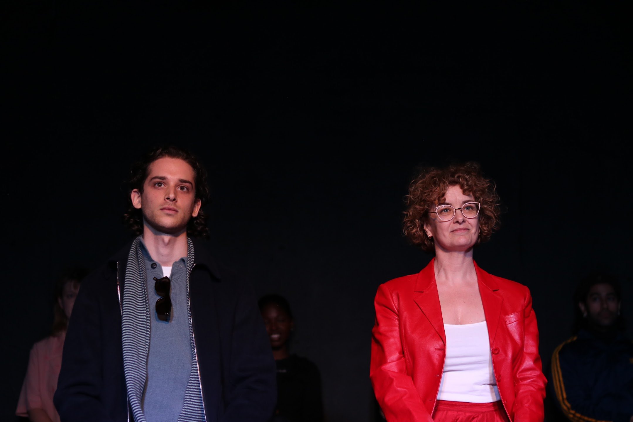 A young man with dark, curly hair wearing a dark jacket, gray sweater, and striped shirt, standing next to an older woman with curly hair and glasses, dressed in a red blazer and white top, against a dark background.