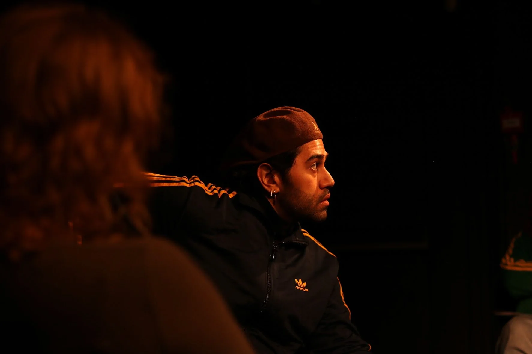 A man wearing a brown beanie and a black Adidas jacket with orange stripes sitting in a dimly lit environment, with a dark background.