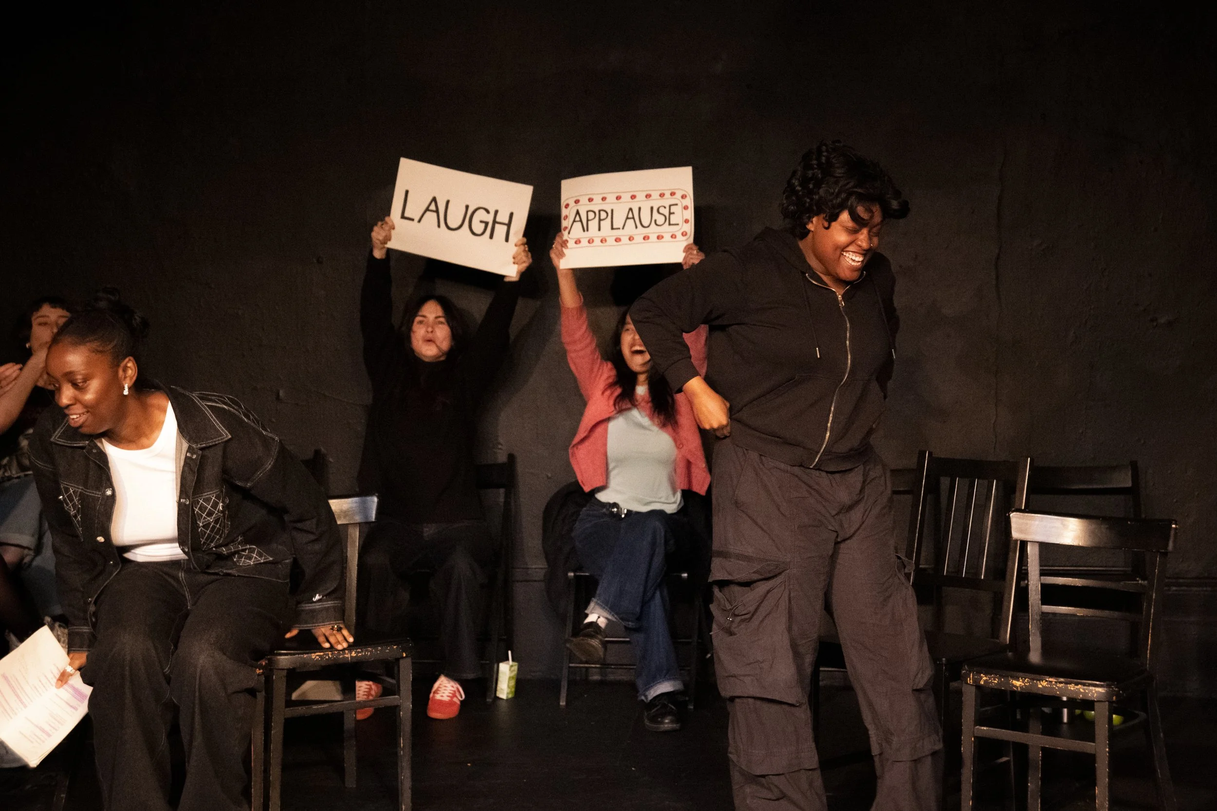 A group of people on stage performing a comedy or theater sketch, with some holding signs that say "LAUGH" and "APPLAUSE." One person is in the foreground, smiling and looking down, while others are seated or standing behind. The background is a blac