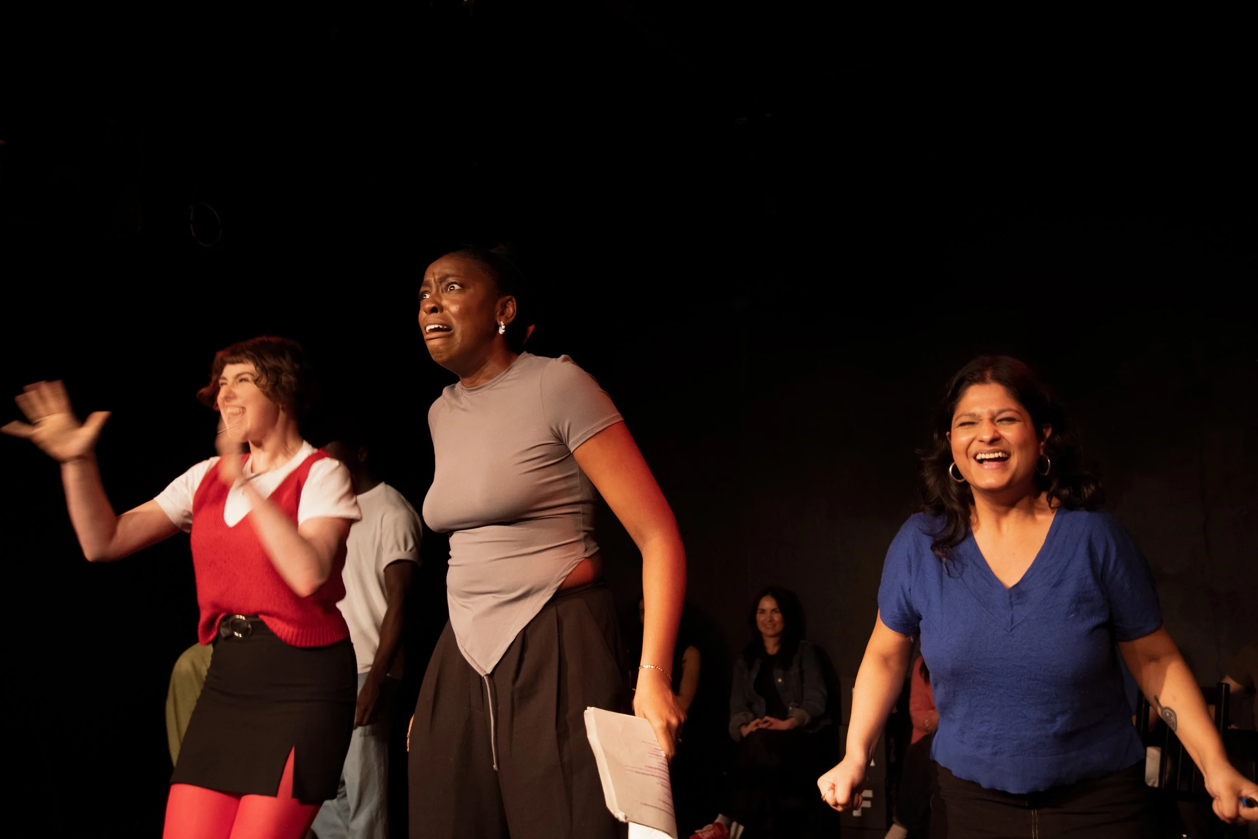 Group of women on stage, some smiling and others with surprised or intense expressions, participating in a performance or event.