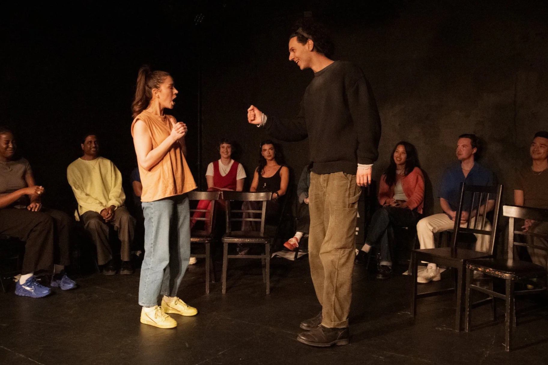 Two actors performing a scene on stage, a woman with a ponytail wearing yellow sneakers and a sleeveless top, and a man with curly hair wearing a black sweater, surrounded by seated audience members watching with various expressions.